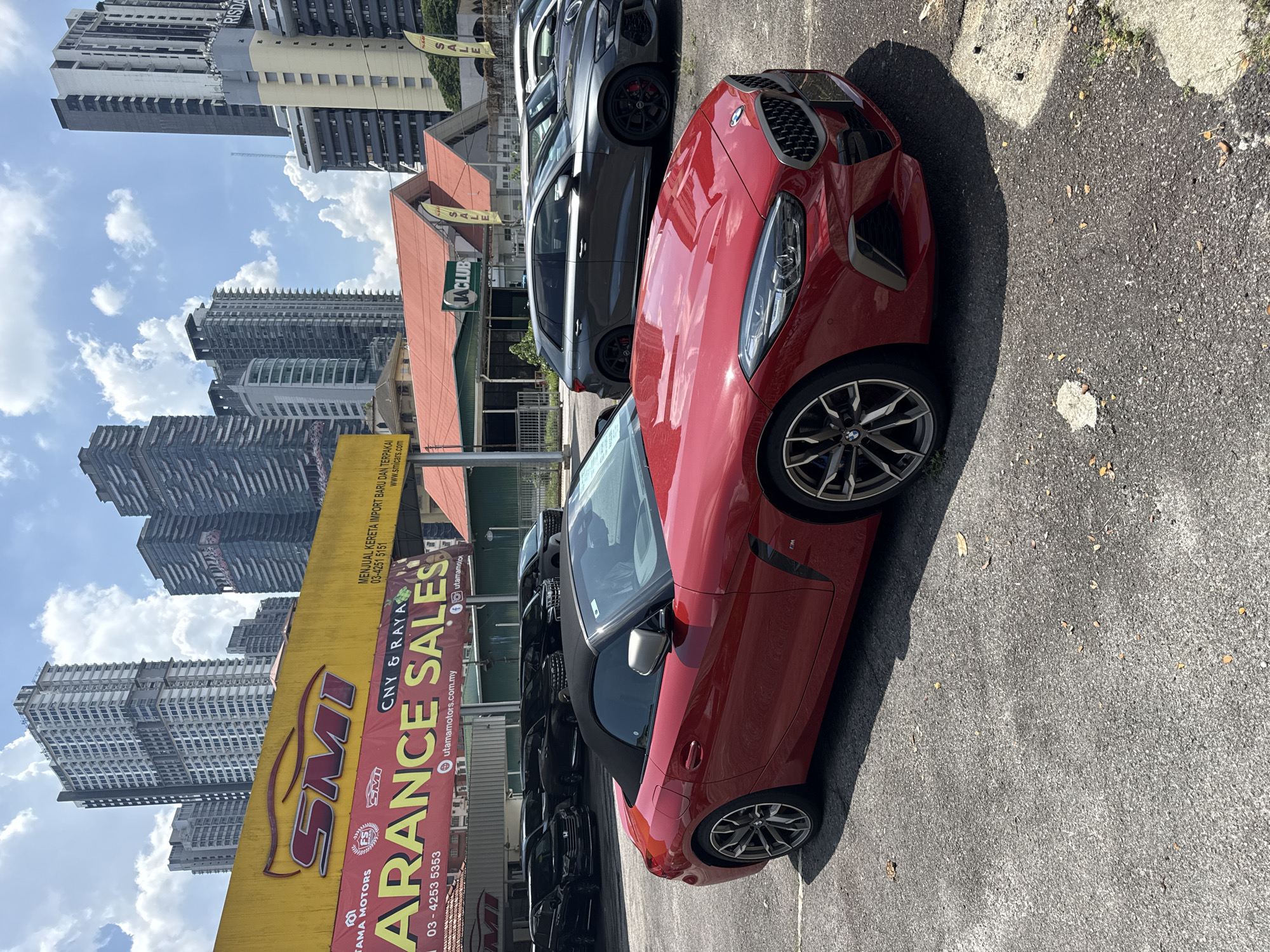 The image shows a red BMW Z4 parked in a lot with other vehicles, possibly at a car dealership in Kuala Lumpur, Malaysia. A large yellow sign for "SMI Utama Motors" advertises "Clearance Sales" and includes contact information. Tall buildings are visible in the background, suggesting an urban setting. The sky is partially cloudy. The photograph is taken during daylight hours.