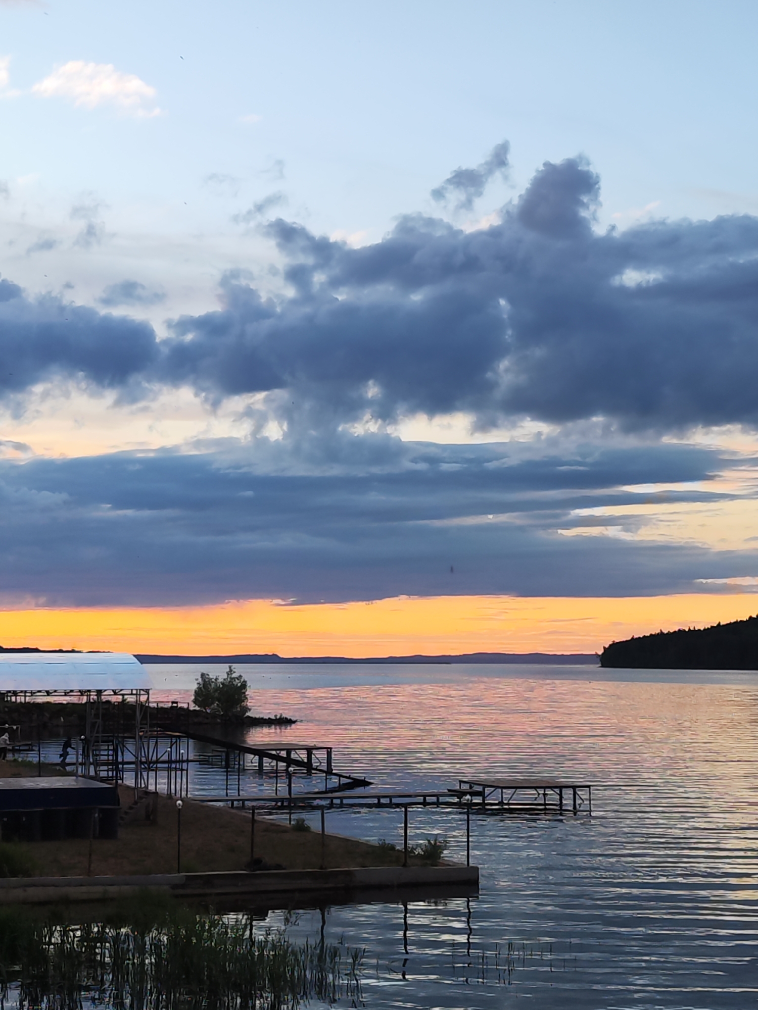 The image shows a sunset over the Kama River in Chaikovsky, Russia. The foreground features the shoreline with wooden piers and structures. Reeds grow in the water near the edge. In the middle ground, the river reflects the colors of the sunset, with hues of orange and pink. Across the river, the opposite shore is visible, marked by a treeline. The sky above is filled with clouds, ranging from light blues to dark grays, reflecting the fading daylight.