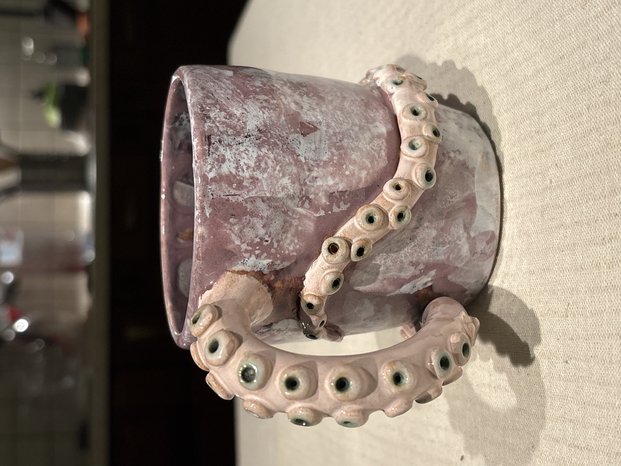 The image depicts a light purple ceramic mug with a handle shaped like an octopus tentacle, featuring visible suction cups. The mug is positioned on a textured, light-colored surface, likely fabric. The background includes a blurred glimpse of what appears to be a kitchen area with cabinets and appliances. There is no indication of the city of Krasnoyarsk, Russia, in the image.