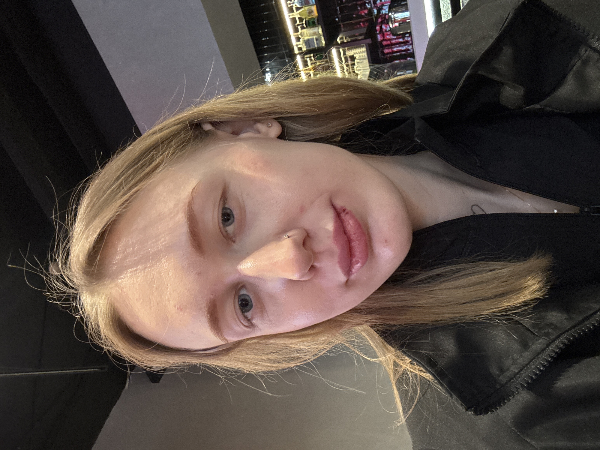A close-up, slightly angled portrait of a blonde woman with fair skin. She has blue eyes and a nose piercing. She is wearing a black jacket. The background appears to be a dimly lit interior, possibly a bar or restaurant with some shelves and bottles visible. There's no definitive evidence to place the image specifically in Rostov-on-Don, Russia.