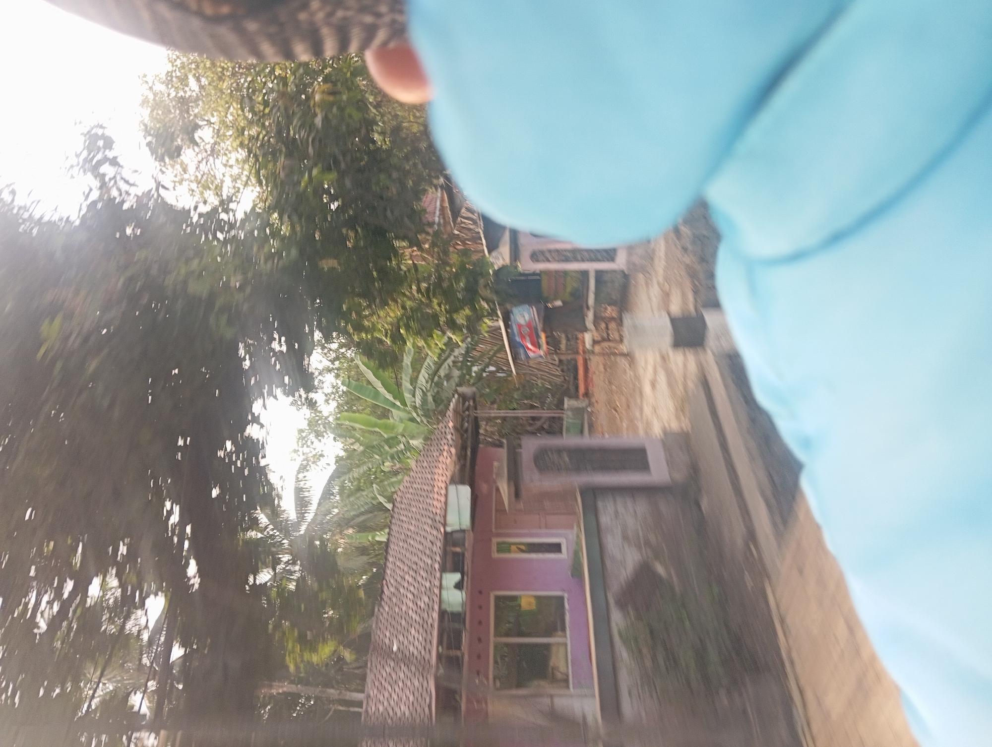 The image shows a side view of a pink house with a tiled roof, located in Klakah, Indonesia. The house has several windows and a light brown door. In front of the house is a low wall. Behind the house, there are trees and other vegetation, including banana trees. The foreground of the image is dominated by the blurred back of a person wearing a light blue garment. The person's head, covered by a patterned cap, is visible on the right side of the frame. The scene appears to be outdoors, with sunlight filtering through the trees. The image has a shallow depth of field, with the background and foreground out of focus.