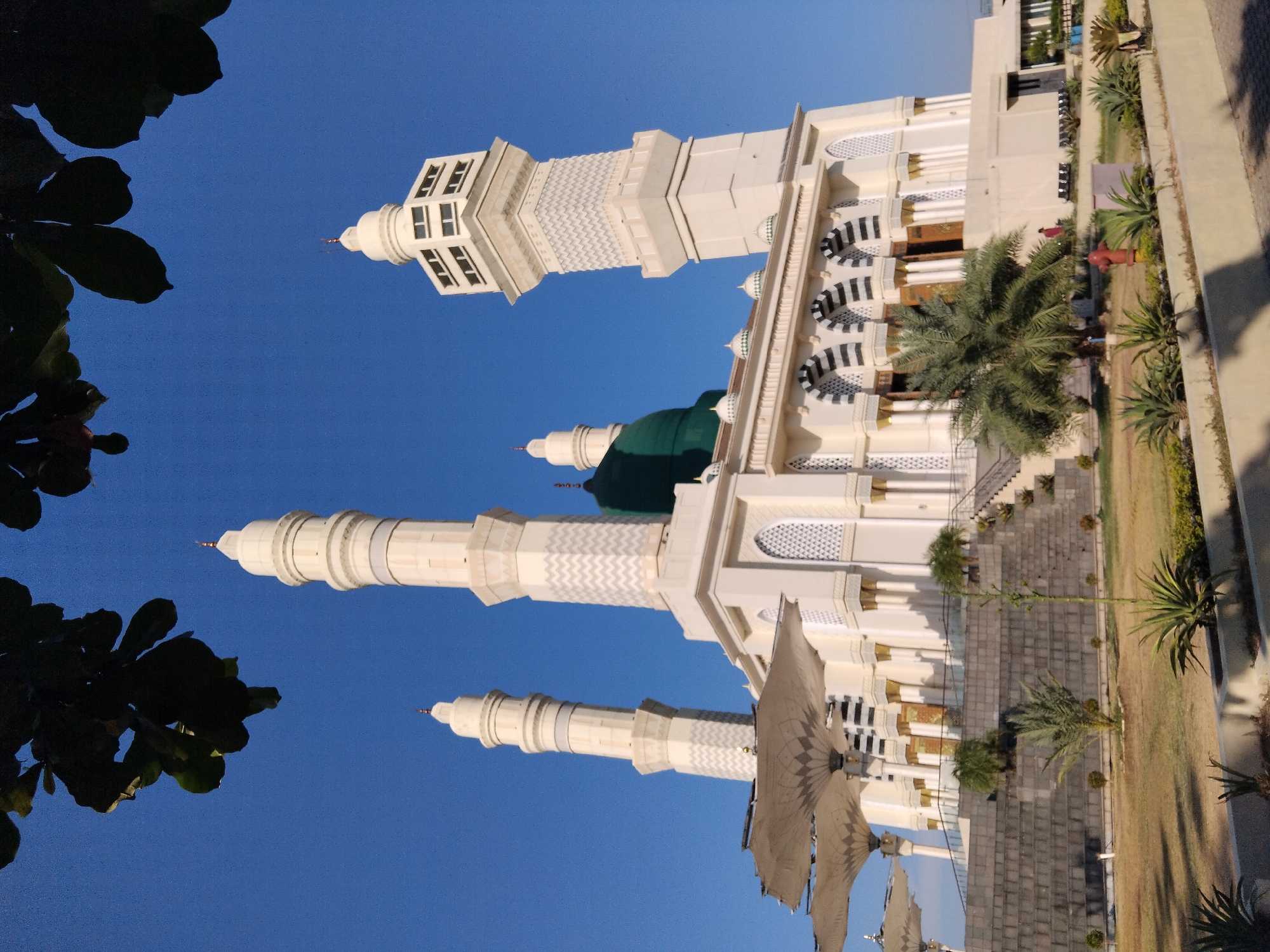 A low-angle, full-shot image captures a large, white mosque with a green dome against a clear blue sky. The mosque features multiple minarets, intricate architectural details like patterned windows and arched doorways, and large, umbrella-like shades in the foreground. A few individuals are visible in the distance, walking or standing. The surrounding area includes a paved pathway, manicured lawns, trees, and low-lying plants. The scene appears to be taken during daylight hours, with shadows indicating the sun is positioned to the side. This mosque is identified as Masjid Agung Madaniyah in Karanganyar, Indonesia.