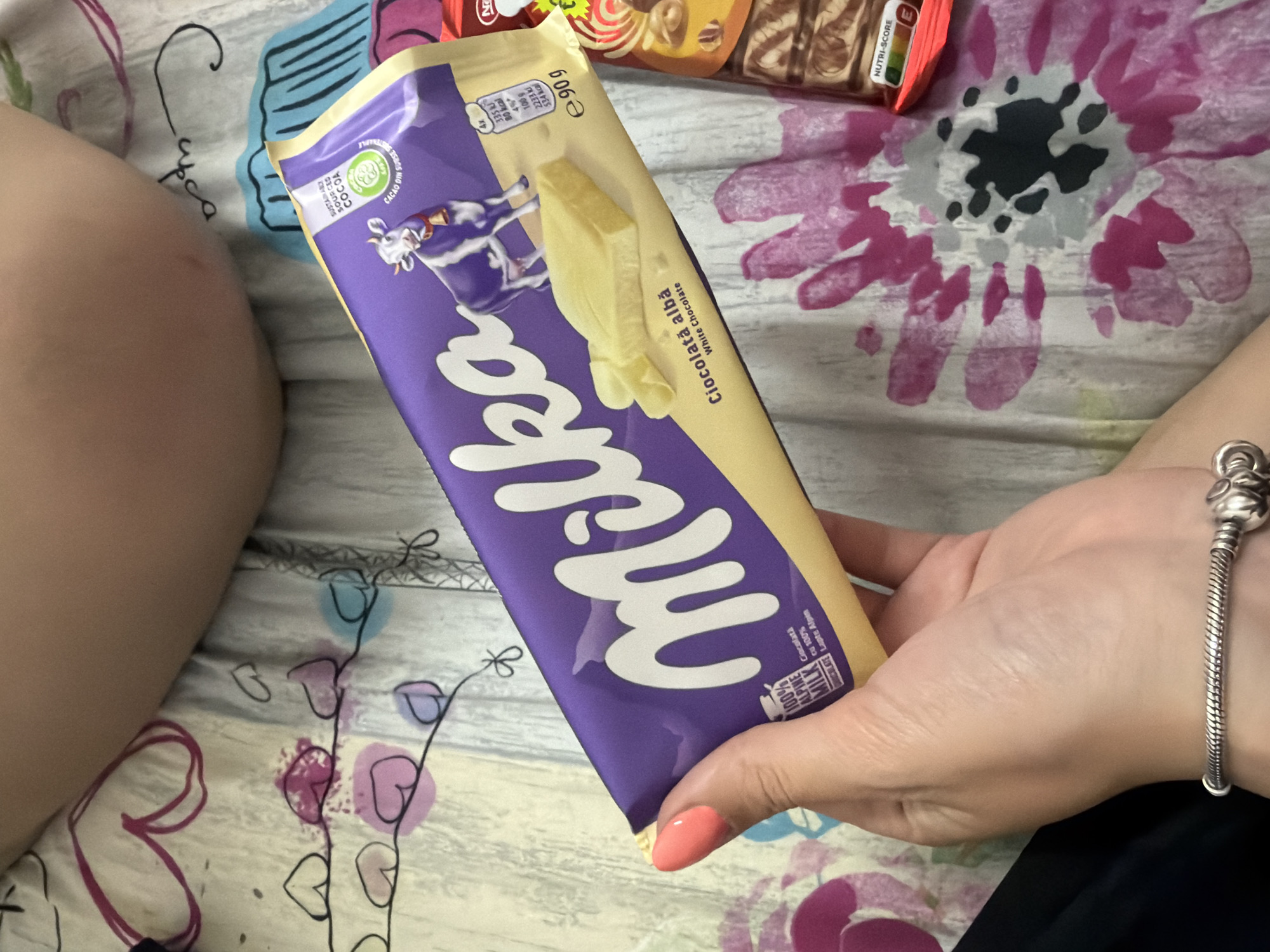 The image shows a person holding a Milka white chocolate bar. The packaging is purple and white with the Milka logo prominently displayed. The label reads "Ciocolata alba" which means "white chocolate". The person is also wearing a silver charm bracelet. The background is a patterned blanket. The city of Sofia, Bulgaria is not discernable in the image.