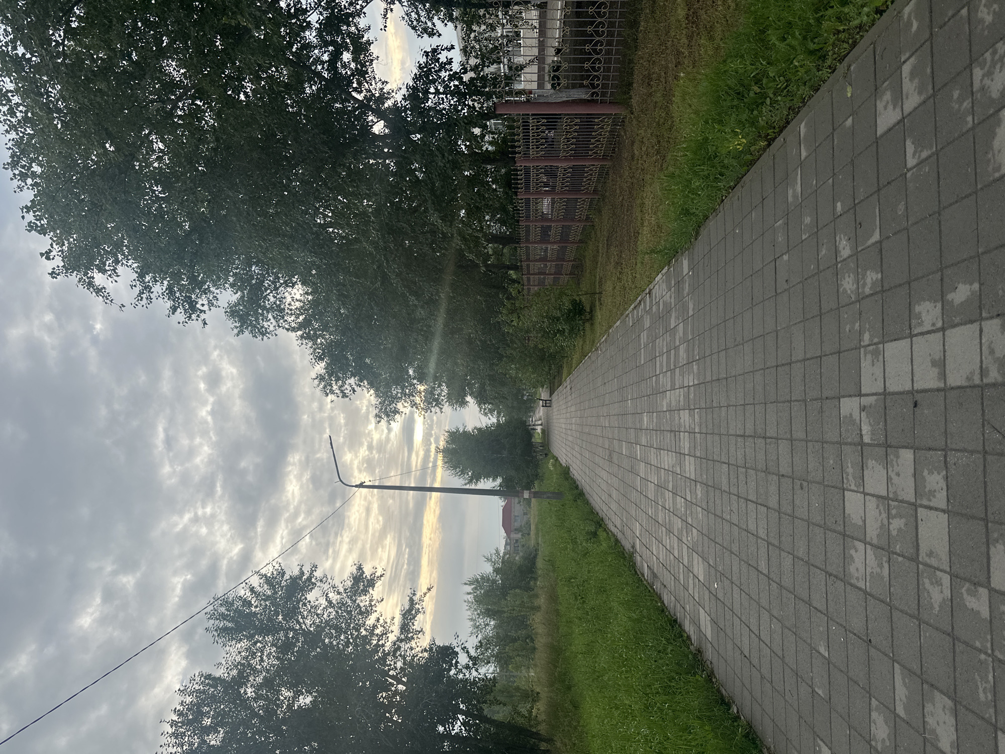 A straight, gray paved walkway, wet in patches, extends into the distance, flanked by a grassy area on the left and a decorative metal fence and trees on the right. A single street lamp is visible near the center of the frame, and further down the walkway, a bench can be seen. In the background, there are buildings and more trees under a cloudy sky. The scene appears to be in Rudny, Kazakhstan.