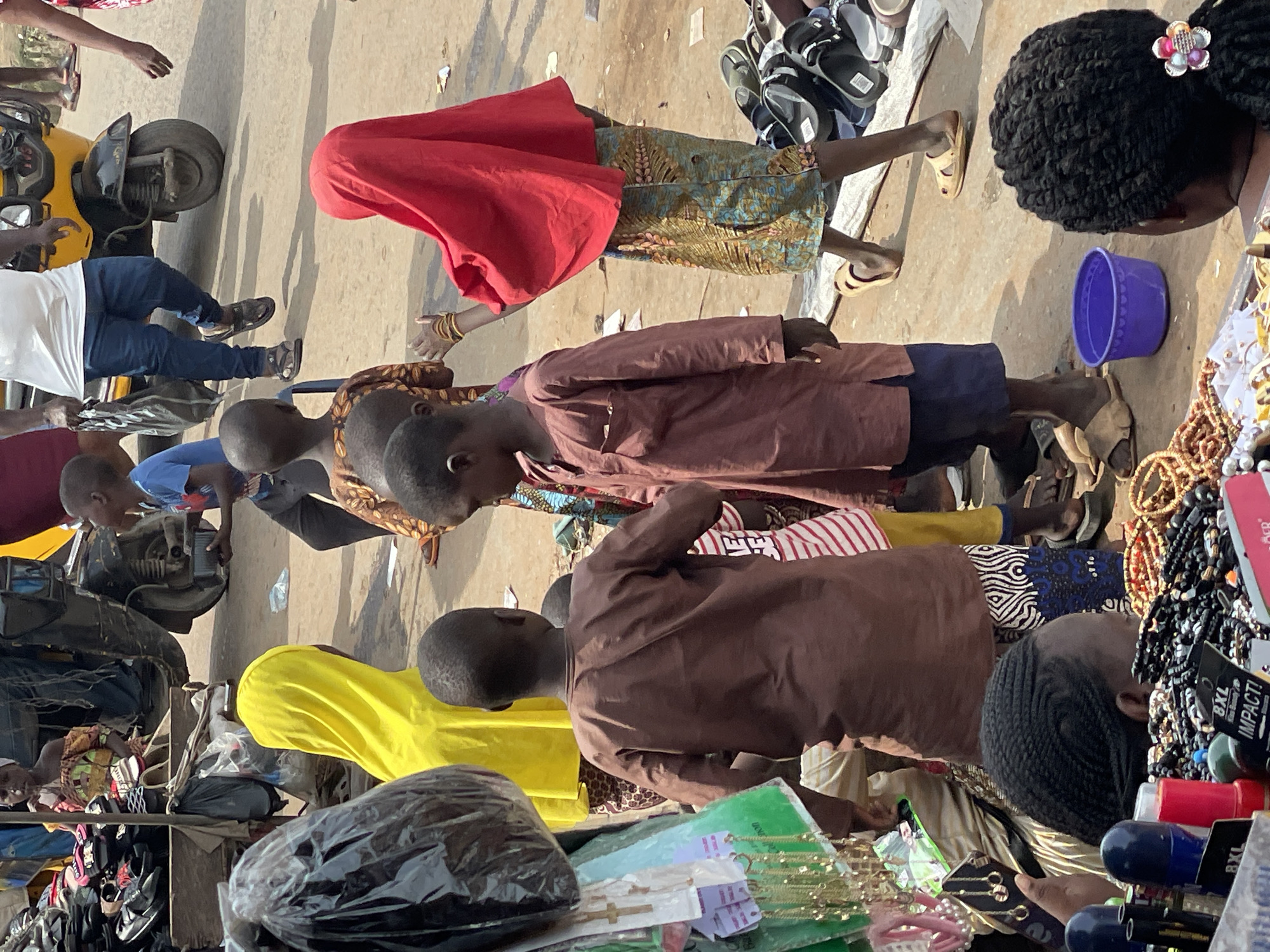 The vertically oriented image depicts a bustling outdoor market scene, likely in a sunny daytime setting. Numerous people, including adults and children, are present, engaging in various market activities.
On the far left, a person in a white t-shirt and blue jeans sits on a yellow and black motorcycle or scooter. Several children and adults are seen standing and walking throughout the scene. A person draped in a bright red headscarf and a patterned skirt is prominent in the upper center, with gold bangles visible on their wrist. Below them, a child in a brown tunic is looking downwards, while another child in a blue t-shirt appears near a metallic object on the ground. A person with a yellow head covering is visible in the lower-left quadrant.
The market stalls are laden with various goods. On the right side, an extensive display of jewelry, including numerous beaded necklaces, bracelets, and earrings, is visible. A purple plastic bucket rests on the ground nearby. A person with intricate braided hair adorned with a flower clip is seen from above, looking down at the merchandise. Another individual with braided hair is also engrossed in viewing items. Stacks of shoes and sandals are visible on shelves or mats in the background. In the foreground and lower-center, items wrapped in black plastic bags, along with other packaged goods, are displayed. Small wooden crosses are also part of the merchandise.
Visible text includes "8XL IMPACT!" and "BXL" on product packaging. The words "LIVE YOUR" are partially legible on a striped shirt. The ground appears to be a dusty or paved surface, suggesting a vibrant public space.