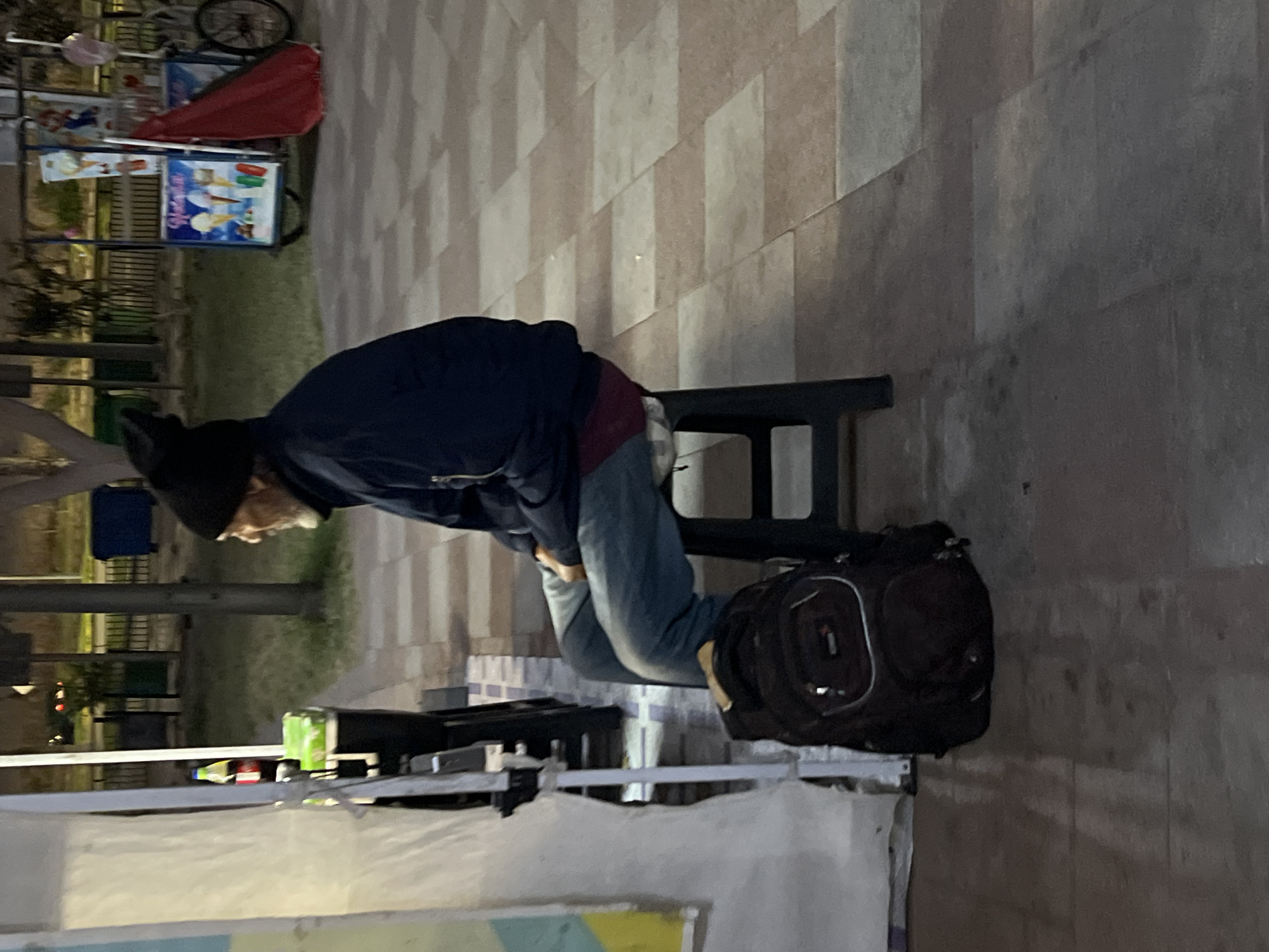 A man in a dark hat and blue jacket sits on a black stool on a stone-tiled surface, with a black backpack resting on the ground beside the stool. A table covered with a white cloth is partially visible in the foreground. In the background, there is an ice cream cart. The scene is Arica, Chile.