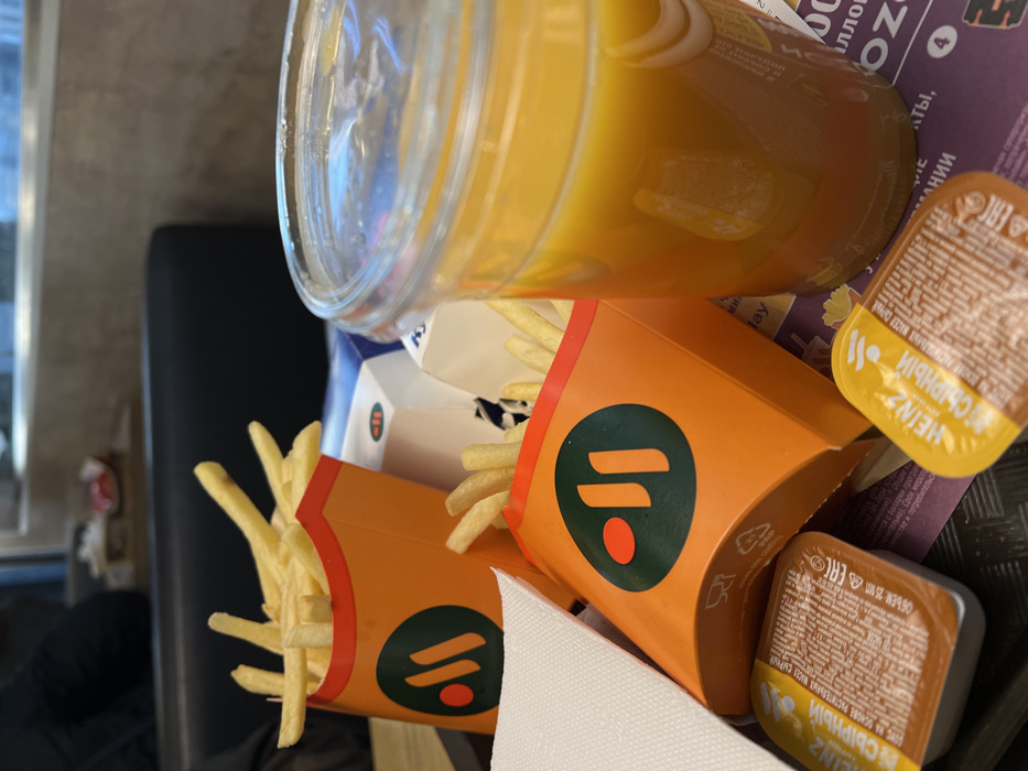 The image displays a close-up, elevated view of a fast-food meal served on a dark brown tray within a "Vkusno i Tochka" fast-food restaurant in Moscow, Russia. The branding on the fry containers and other packaging, along with Cyrillic text on labels, confirms the location as a "Vkusno i Tochka" establishment.
On the tray, two orange cardboard containers filled with golden-brown french fries are prominently featured. Each container bears the green "Вкусно и точка" logo. To the right, a clear plastic cup with a lid holds an orange-colored beverage, likely juice. At the bottom left, a folded white paper napkin is visible. Below the right fry container and next to the beverage, two small brown Heinz sauce packets with yellow labels, marked "СЫРНЫЙ" (Cheesy) in Cyrillic, rest on the tray. Partially visible behind the front fry container is a white and blue cardboard box, likely containing a burger, with some Cyrillic text visible.
The background is softly blurred, showing the interior of the restaurant. A dark upholstered booth seat is visible, and behind it, a person wearing dark clothing is partially obscured, seated at a table or booth. The overall environment suggests a typical indoor fast-food dining area with artificial lighting.