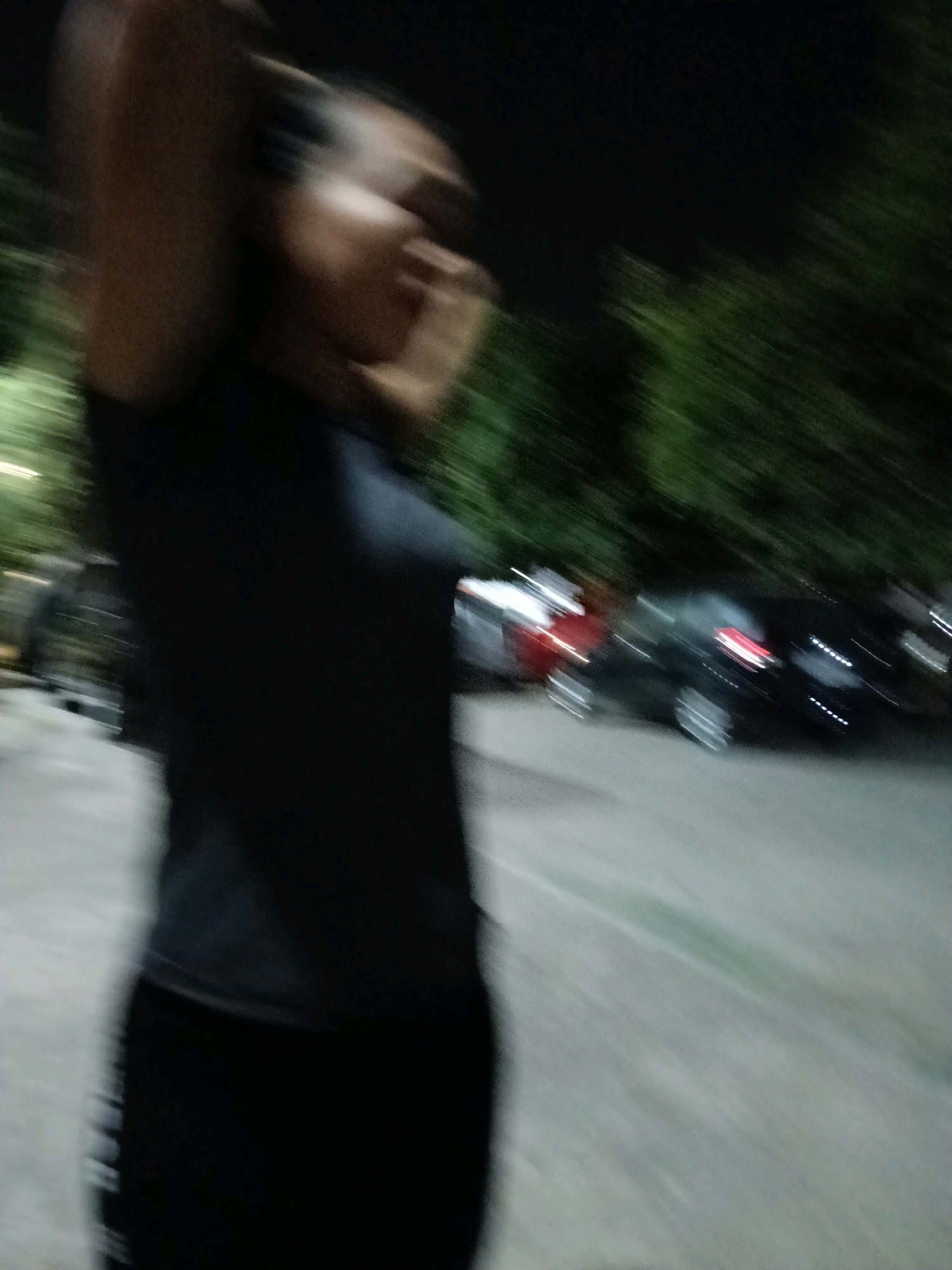 The image captures a blurred motion of a person in a dark shirt and pants, standing in what appears to be an outdoor parking lot at night. The person's arms are raised above their head, and they are facing to the left of the frame. In the background, several cars are visible, including what looks like a dark-colored sedan with its brake lights illuminated. Trees line the background, suggesting an urban or suburban setting. The ground is paved, likely asphalt or concrete. The lighting is dim, consistent with nighttime conditions, with some artificial light sources possibly contributing to the illumination. The location is Jatimulya, Indonesia.