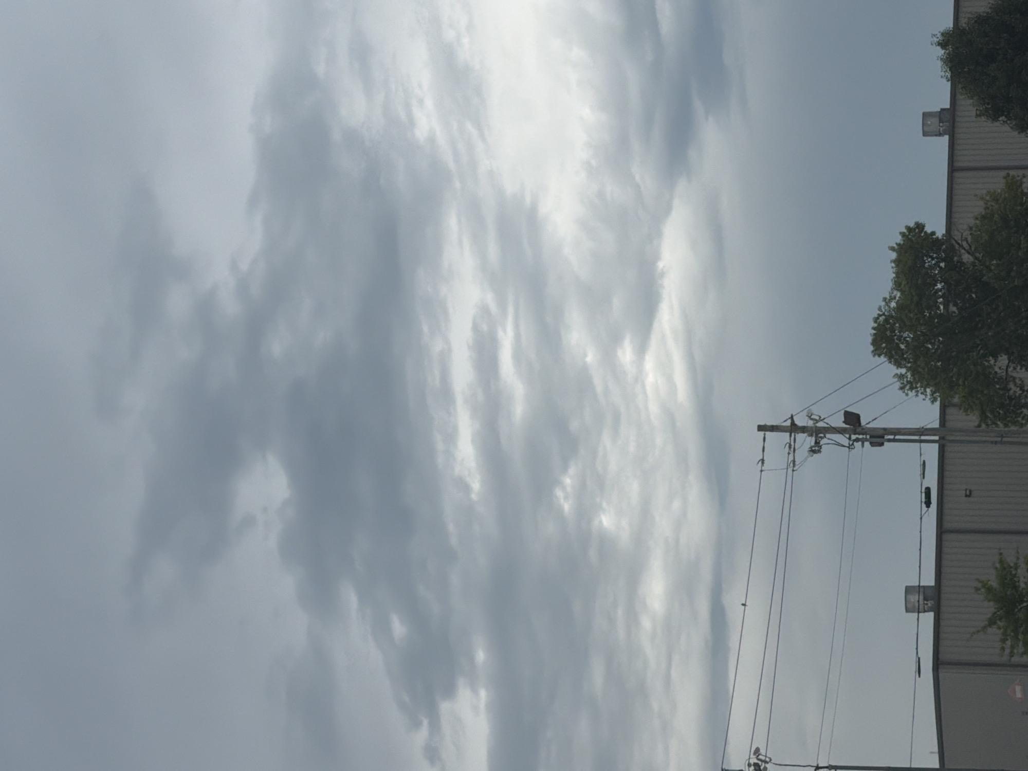 The image shows a cloudy sky over a section of Murfreesboro, United States of America. In the foreground, a grey, industrial-style building with a corrugated metal facade is visible. On top of the building, there are two cylindrical vents. Utility poles with multiple wires and streetlights run horizontally across the frame above the building. A few trees with green foliage are positioned in front of and to the side of the building. The sky is overcast with varying shades of grey clouds.