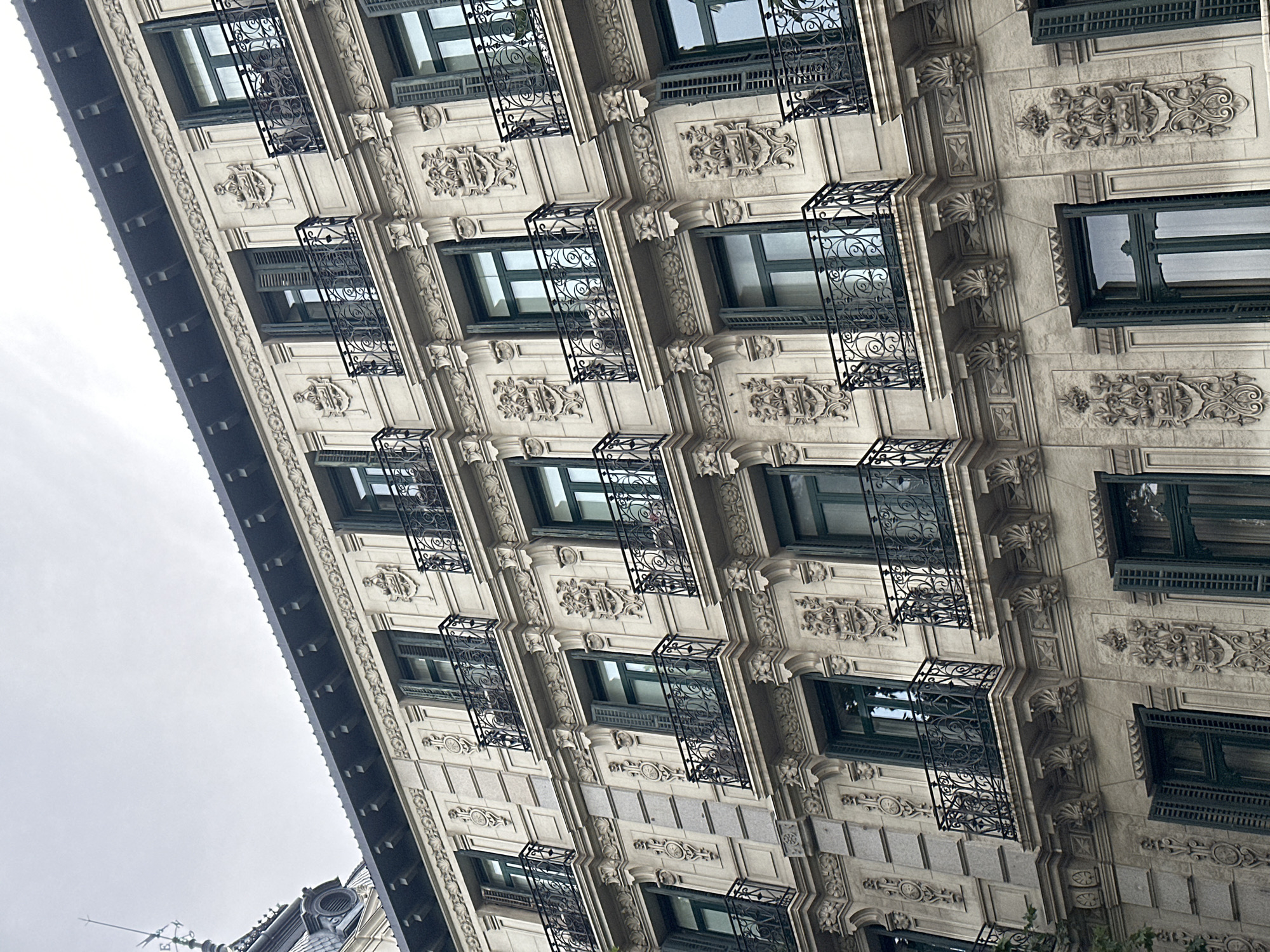 The image depicts the exterior of a multi-story building in Madrid, Spain. The building is characterized by an ornate, light-colored facade with intricate architectural details, including decorative carvings and moldings. The windows are framed in dark green and some feature decorative black iron balconies. The building is seen from a low angle, partially obscured by an overcast sky. A glimpse of a neighboring structure with a spire is visible at the bottom left.