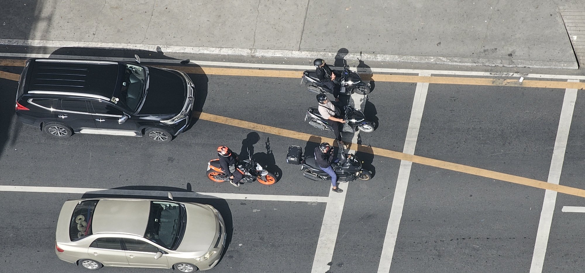 This is an aerial, high-angle shot of traffic on an urban street in Makati City, Philippines. The scene is captured during daylight, as evidenced by the distinct shadows cast by the vehicles and people. The road surface is asphalt, marked with white and yellow traffic lines.
In the foreground, a silver sedan is parked or stopped, facing right. The car's interior is visible, revealing items stacked on the rear seat and in the trunk area. To the left of the sedan and slightly further up the road, a black SUV is in motion, moving towards the right side of the frame.
The middle ground and background feature several motorcycles and their riders. One orange and black motorcycle, with a rider wearing a red helmet and dark clothing, is positioned near the silver sedan. Further up the road, two black scooters are stationary, each with a rider wearing a helmet. One rider, in a light-colored shirt, is looking towards the right. Another black scooter is also stopped, with a rider in dark clothing wearing a black helmet.
The environment includes a concrete sidewalk bordering the road on the left, with a faint shadow of a structure or tree. The overall scene depicts a typical urban traffic condition with vehicles and motorcyclists navigating the street.