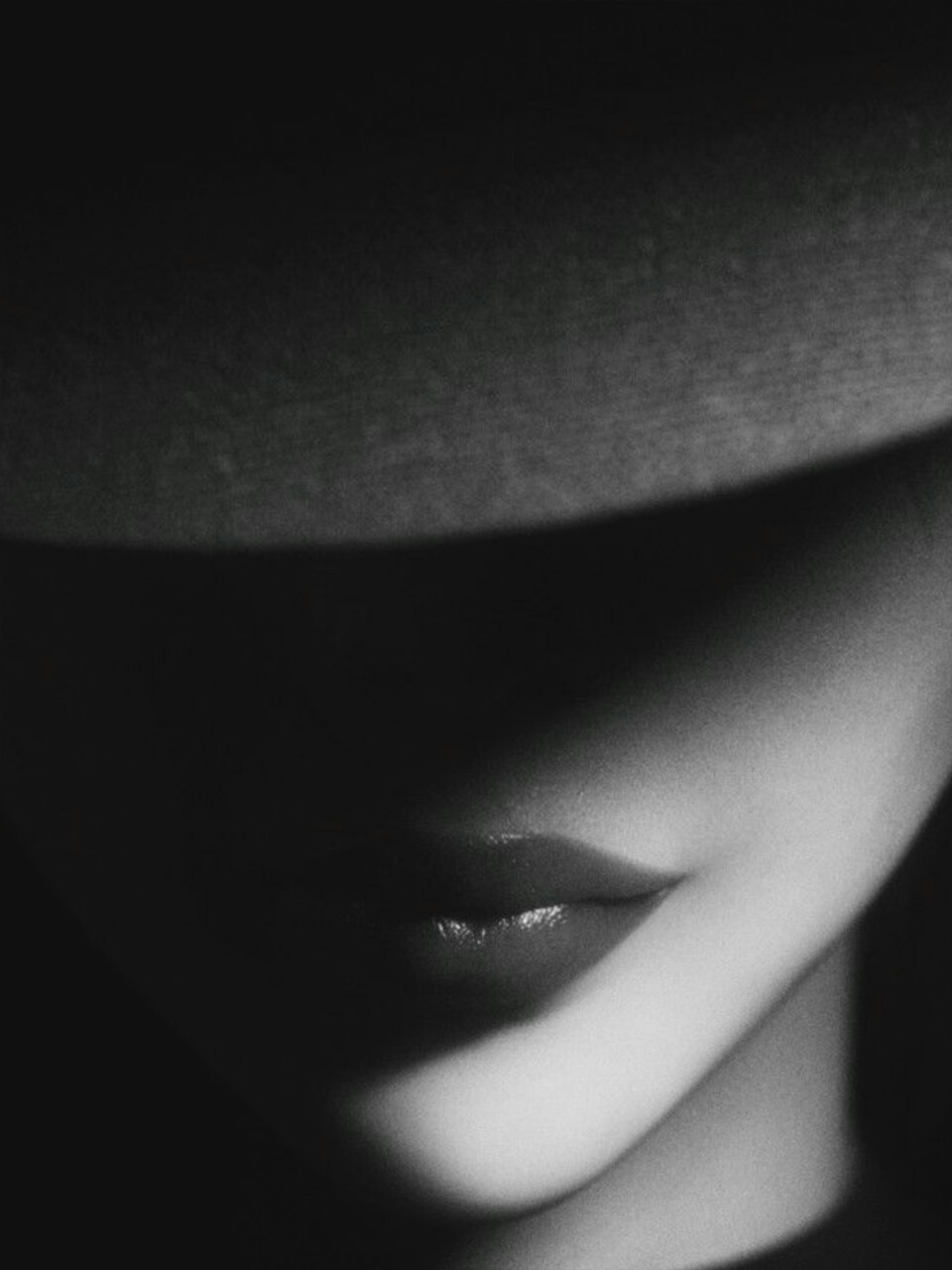 The image is a close-up, low-angle, black and white shot of a person's mouth and chin, partially obscured by shadow cast by a hat. The lips are slightly parted and appear to be wearing lipstick. The lighting creates a dramatic contrast between the shadowed areas and the highlighted skin. The background is dark and indistinct, with no discernible environmental features or urban elements. There is no indication of the location being French Pass / Anaru, New Zealand.
