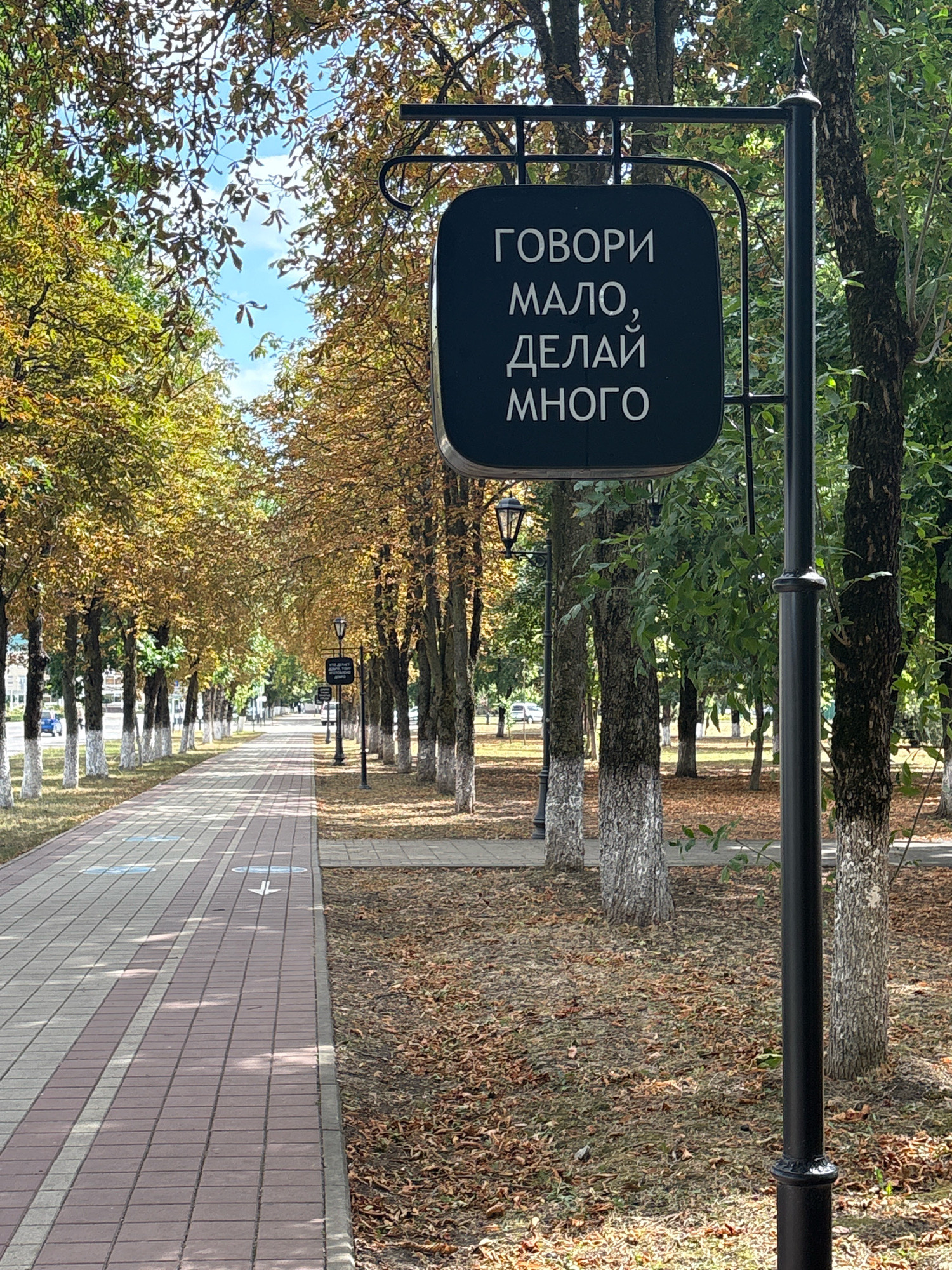 The image depicts a tree-lined pathway in a park, likely in Maykop, Russia, judging by the text on the sign. The pathway is paved with brick-like tiles in alternating shades of pink and grey. To the left of the pathway, trees with yellowing leaves, indicative of autumn, line the area. To the right, the ground is covered with fallen leaves. A lamppost stands prominently in the foreground, to the right of the pathway. Attached to the lamppost is a black, oval-shaped sign with white Cyrillic text. The text on the sign reads: "ГОВОРИ МАЛО, ДЕЛАЙ МНОГО" which translates to "Speak little, do much." The sky is visible through the canopy of the trees, with some clouds present. Further down the pathway, additional lampposts and trees can be seen, creating a sense of depth. There are no people visible in the image.