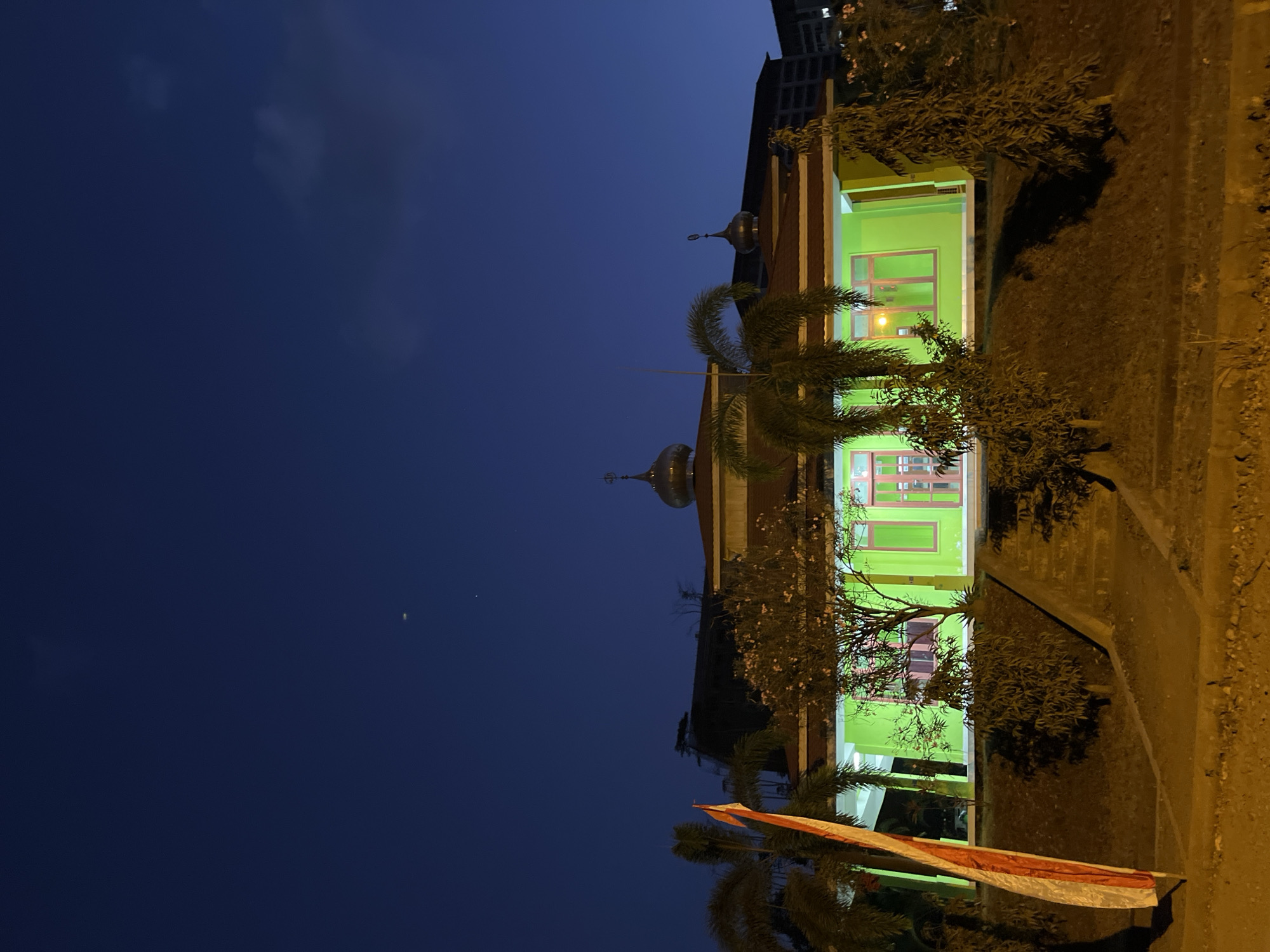 The image shows a mosque in Lalampu, Indonesia, at dusk. The mosque is illuminated with green light, and its domes are visible against the dark blue sky. There are palm trees and other vegetation around the mosque, as well as a flag pole with a red and white flag. In the background, a building is visible. The sky has a faint crescent moon and a few stars. The foreground is a dirt road with streetlights casting an orange glow.