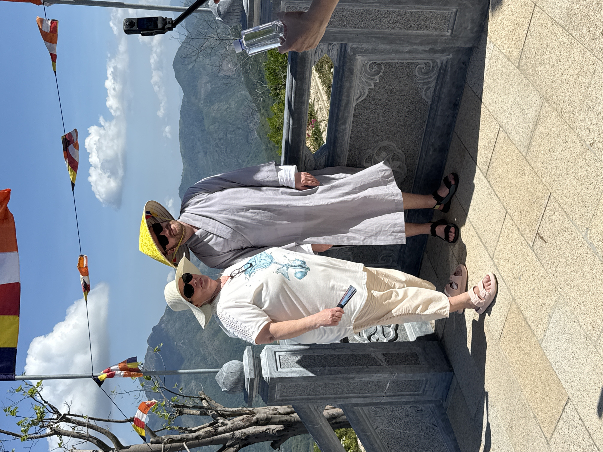 Two individuals are standing on a stone balcony with intricately carved railings in Nha Trang Ward, Vietnam. The backdrop reveals a mountainous landscape under a partly cloudy, blue sky. Buddhist prayer flags are strung up in the foreground.
The person on the left is wearing a white sun hat, sunglasses, a white shirt with a blue design, light beige pants, and pink sandals. They are holding a black folding fan. The person on the right wears a traditional yellow conical hat, sunglasses, and a light gray robe, with black sandals visible on their feet.
An arm holding a clear water bottle can be seen above the person in the gray robe, while a camera mounted on a stick is positioned above the person wearing the yellow hat. The scene appears to be captured in bright sunlight.