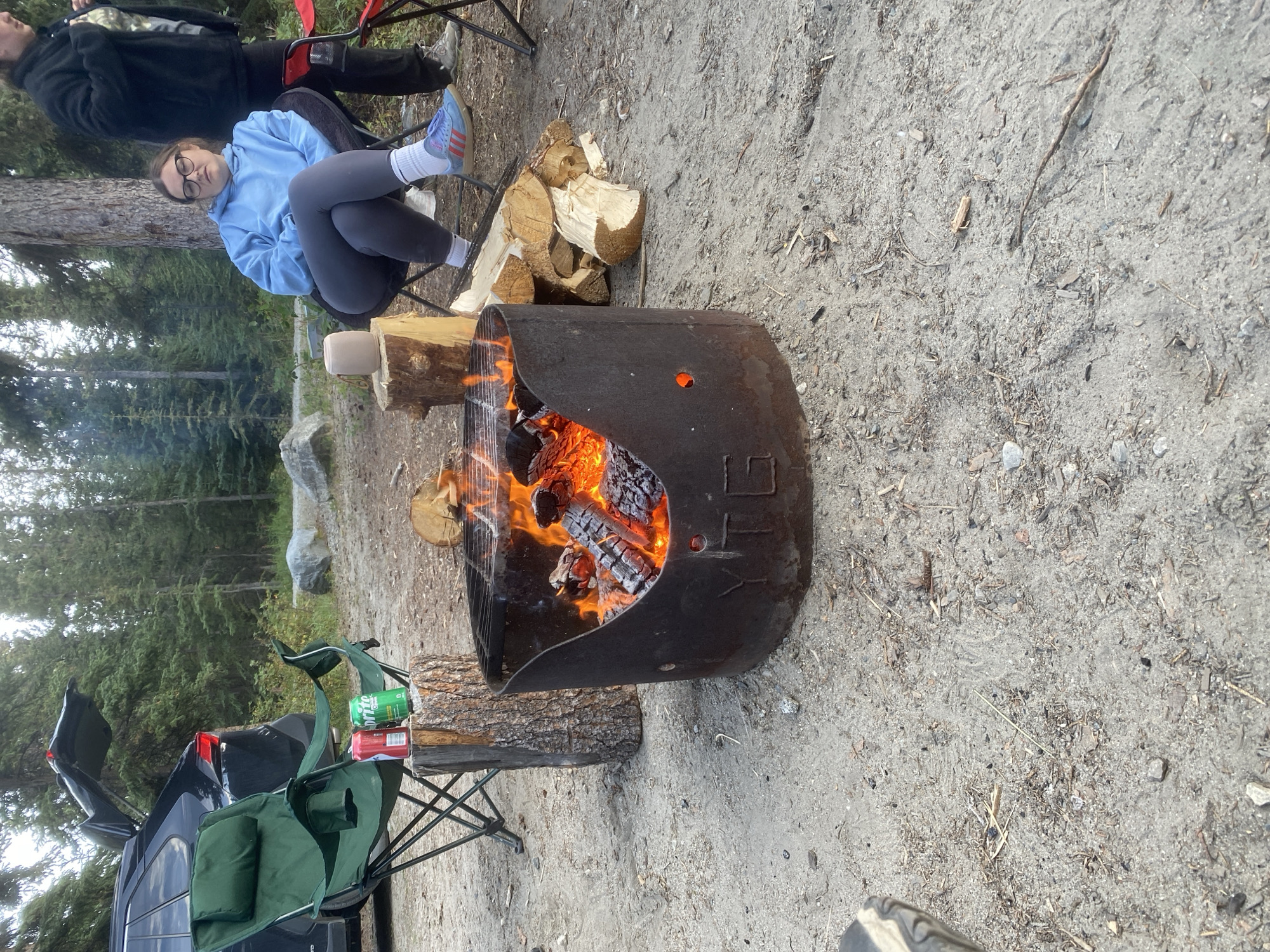 The image depicts a campsite in Haines Junction, Canada, with a campfire at the center. A circular metal fire pit contains burning logs and embers, with a grill on top. To the left, a green camping chair is set up next to a dark-colored SUV with its trunk open. Two beverage cans, one red and one green, rest on a wooden log. In the background, a young woman wearing a blue hoodie and grey leggings is seated in a chair, facing the fire. Another person, partially visible, stands to her right. The ground is sandy and covered with dirt, small rocks, and scattered pine needles and twigs, indicative of a forest environment. The surrounding area is characterized by a dense forest of evergreen trees and scattered large rocks.