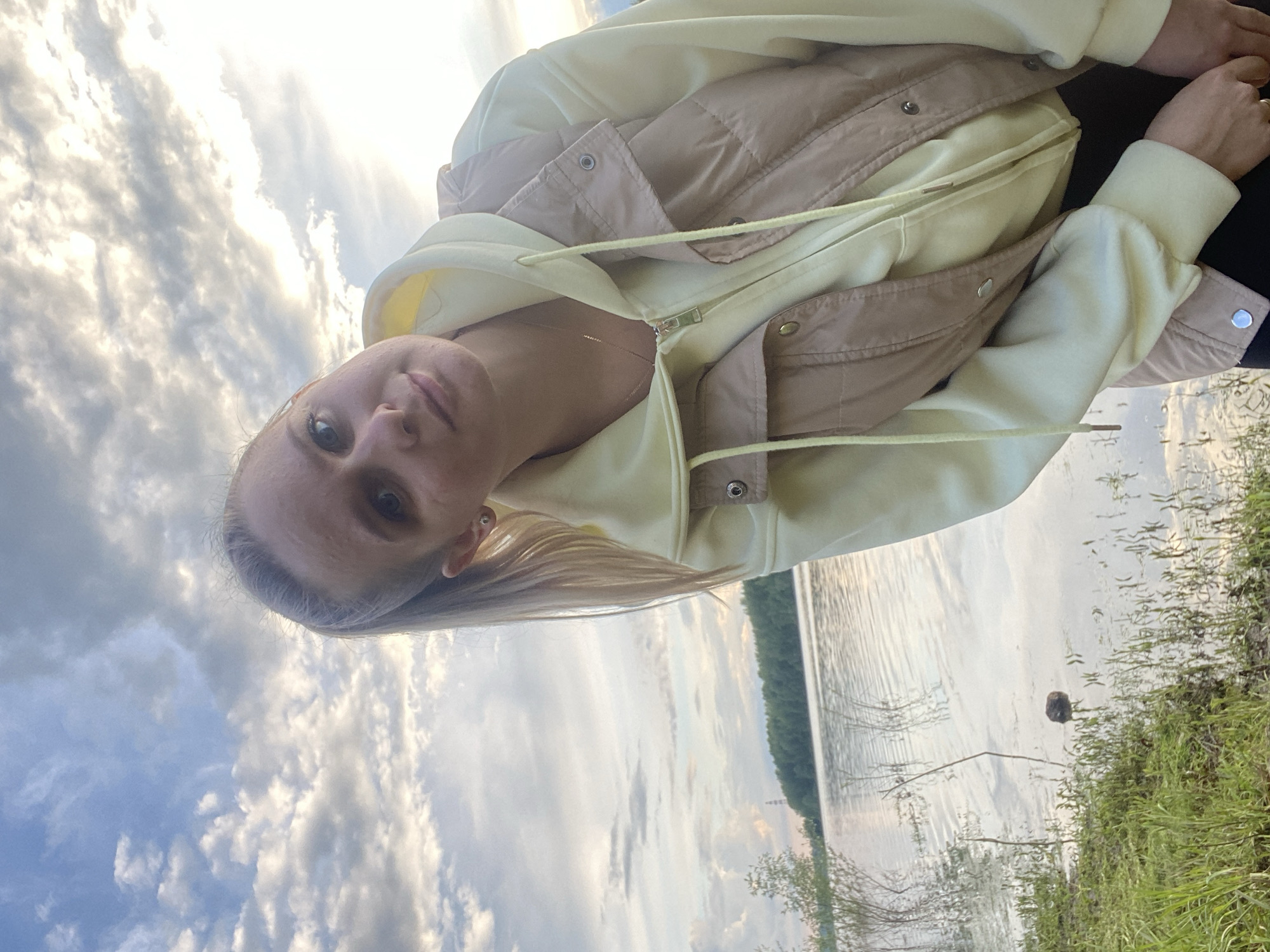 A blonde woman with gray-blue eyes and fair skin stands in front of a body of water, likely the Kama River, in Chaikovsky, Russia. She wears a light yellow hoodie under a tan puffy vest. The sky is partially cloudy. Green foliage is visible near the water's edge. A dark object floats in the river.