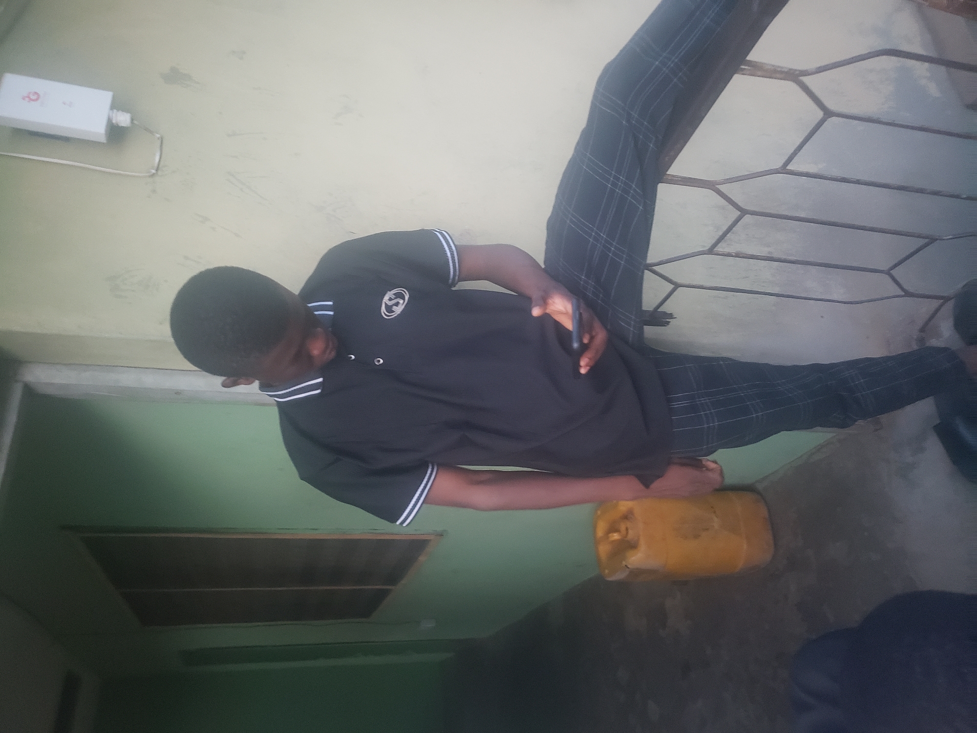 The image shows a person standing near a building in Ibadan, Nigeria. The person is wearing a black polo shirt with white stripes on the sleeves and collar, and plaid pants. They are holding a mobile phone in their hand. A yellow jerrycan is on the ground near the person's feet. A metal security gate and a white box attached to the wall are visible in the background. The building's walls are painted in shades of green and off-white.