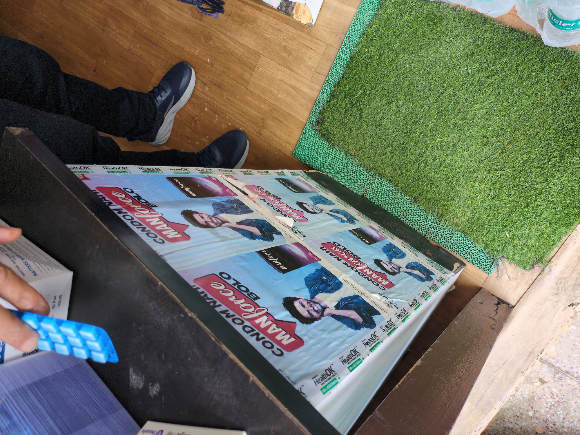 In Bhagsu, India, a close-up shot captures a stall displaying promotional material for "Manforce Bolo" condoms. A hand holding a blue plastic object, possibly a condom dispenser, is visible in the upper left corner. The stall features vibrant posters with images of men and the product branding. In the background, a person's legs clad in dark pants and blue athletic shoes are visible, standing near a wooden floor. A green mat with a textured pattern resembling grass is placed on the floor, adding a touch of nature to the commercial setting. The overall scene suggests a retail environment where personal care products are being marketed.