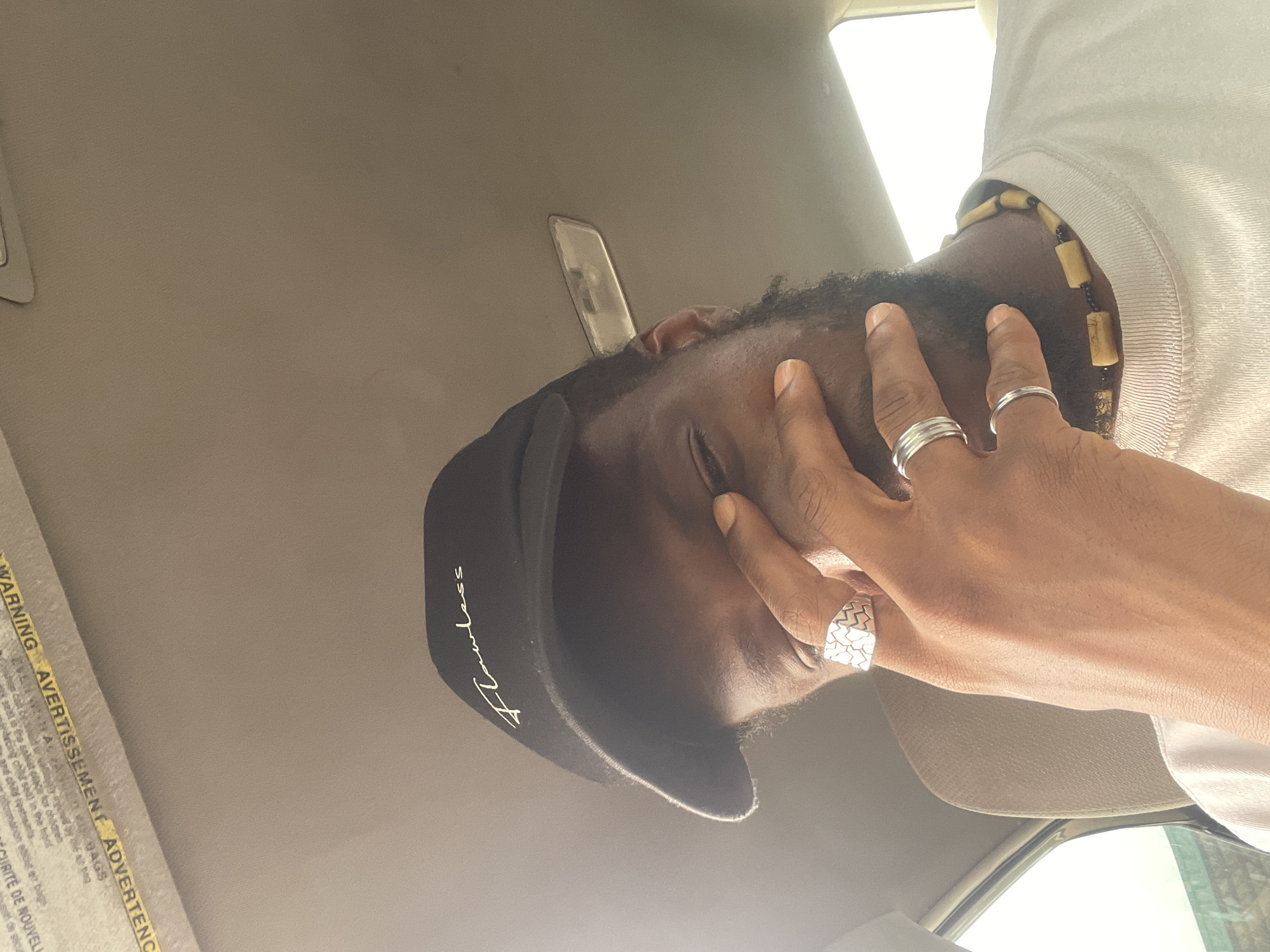 This media file shows a close-up, angled shot of a person's head and hand, with a focus on their face, particularly their closed eyes and the hand covering part of their forehead. The person appears to be wearing a black baseball cap with the word "Flawless" printed in gold lettering. They are also wearing a beaded necklace and two silver rings on their fingers. The background suggests an indoor setting, possibly the interior of a vehicle, with a beige-colored headliner visible. The lighting indicates it might be daytime, with bright light coming from the right side of the frame, possibly a window. There is some visible text on a yellow and white label in the bottom left corner, which appears to be a warning label, with "WARNING", "AVERTISSEMENT", and "ADVERTENCIA" legible. The overall mood conveyed is one of introspection or weariness, with the hand shielding the eyes.