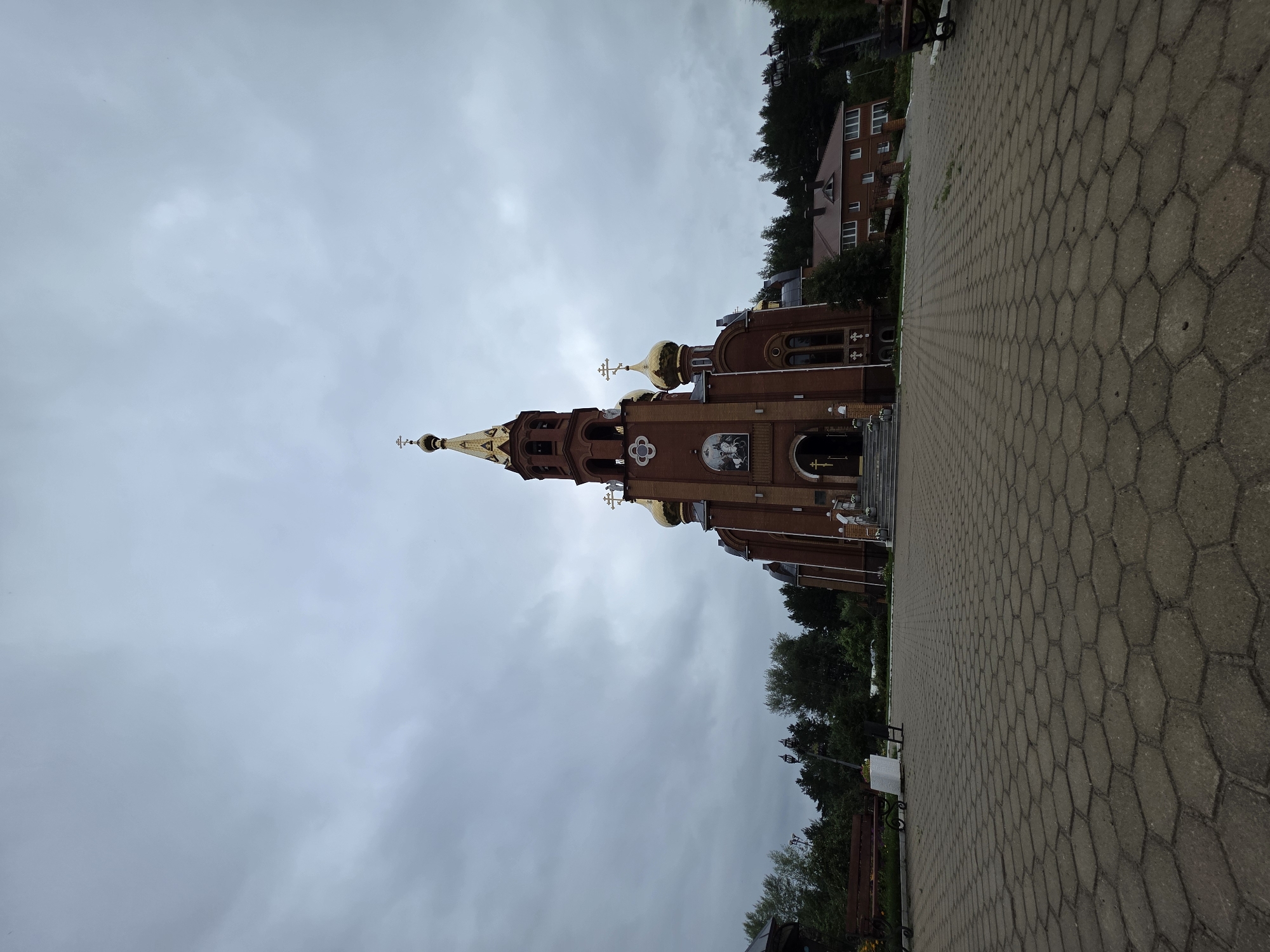 This is a medium-angle, wide-shot, eye-level, outdoor photograph of the Church of the Assumption of the Blessed Virgin Mary in Chaikovsky, Russia, on a cloudy day. The brick church has golden domes and a tall bell tower topped with a golden cross. In front of the church is a large paved courtyard with hexagonal gray paving stones. To the left of the church, there are trees and a park bench. To the right of the church, there is another building and more trees. The sky is overcast and gray.