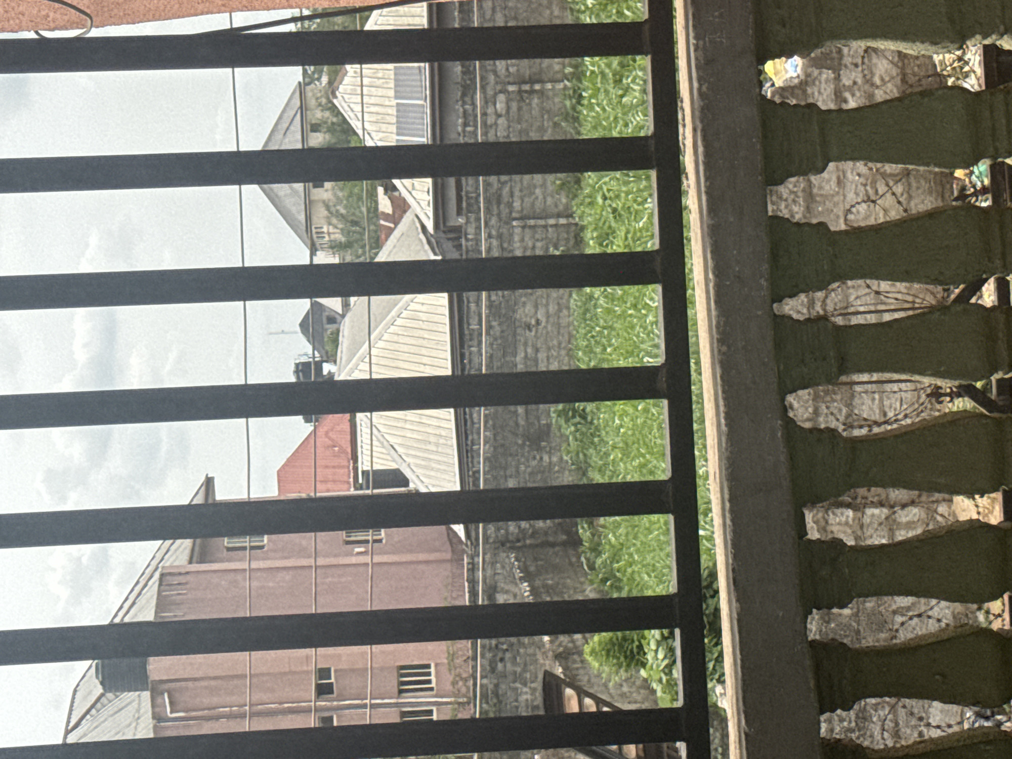 View from behind a metal bar fence and decorative concrete railing, looking out onto a residential area in Ihiagwa, Nigeria. Buildings of varying sizes and colors are visible, interspersed with greenery. One building is a multi-story pink building. A gray stone wall with plants is also present. The sky is overcast. No people are immediately visible.
