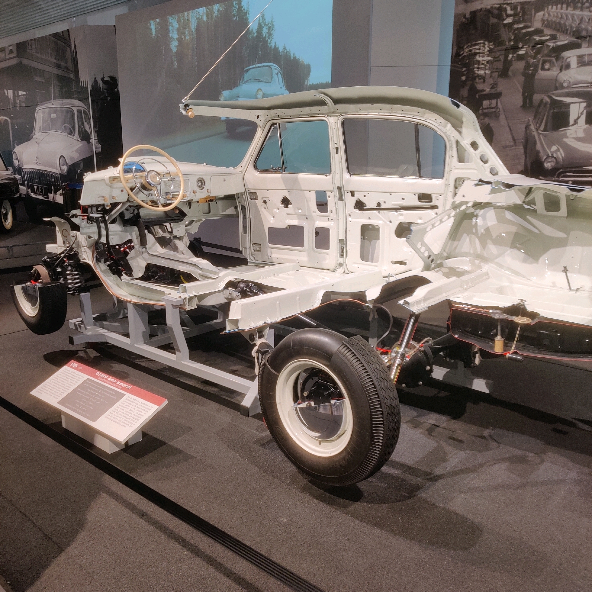 The image depicts the chassis and interior structure of a white car, likely a GAZ-20 "Victory" or a similar Soviet-era model, displayed in a museum. The car's body panels are removed, exposing the frame, engine components, suspension, and interior framework. A steering wheel is visible in the driver's position. In the background, large photographic murals show vintage cars and possibly a factory setting. To the left, a partially visible vintage car with the license plate "XLE 1" is present. In the foreground, a white informational placard with text in Russian and English is displayed. The scene is illuminated by museum lighting, casting shadows on the dark floor. This exhibition is located at the UMMC Museum of Military and Automotive Equipment in Verkhnyaya Pyshma, Russia. No people are visible in the immediate scene.