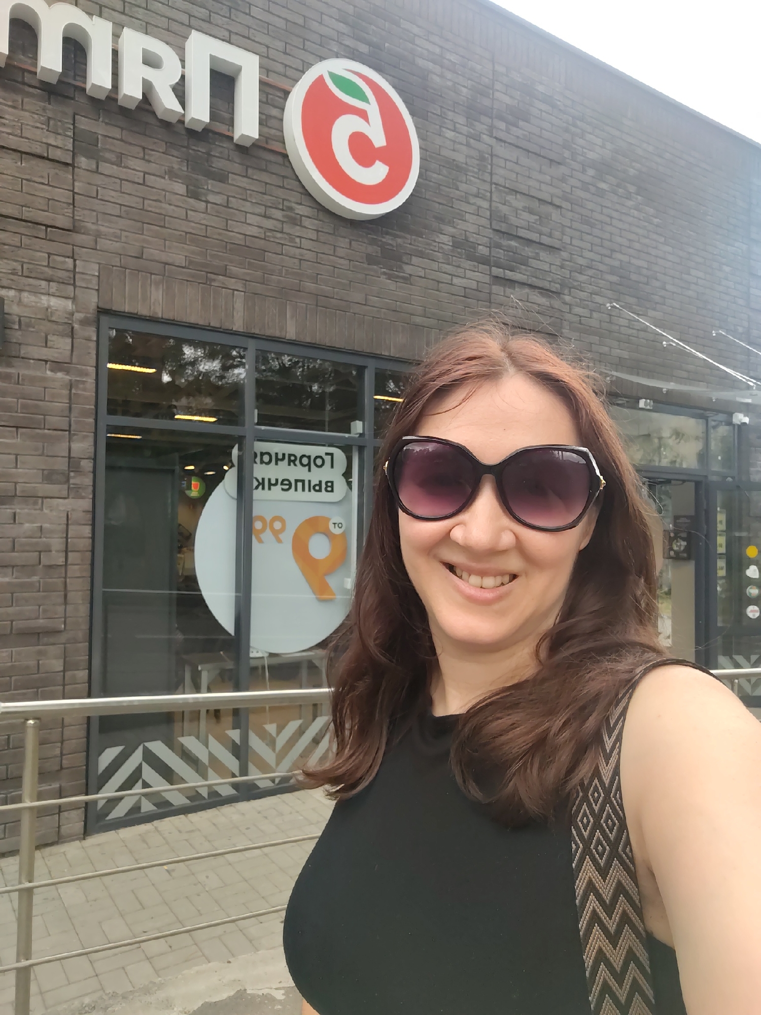 A woman with shoulder-length brown hair and large sunglasses smiles at the camera in front of a brick building. The building has a sign that says "mrn" in large white letters and a red circular logo with a green leaf. In the background, through a large glass window, a sign with Cyrillic text is visible, reading "ЛЮБИМАЯ" (Lyubimaya). The scene appears to be in Rostov-on-Don, Russia, as suggested by the store name which is a common chain in that region.