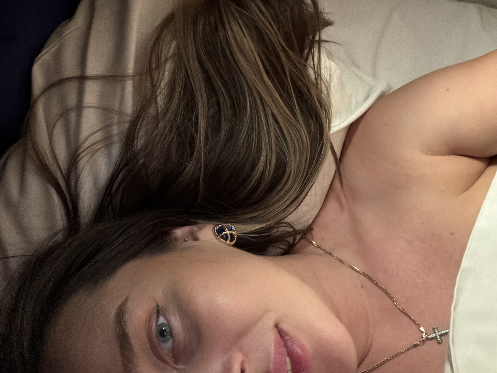 A woman with light brown hair, wearing a blue and gold earring and a gold necklace with a cross pendant, is lying on a bed with a white pillow and sheet. The location is not identifiable as Moscow, Russia.