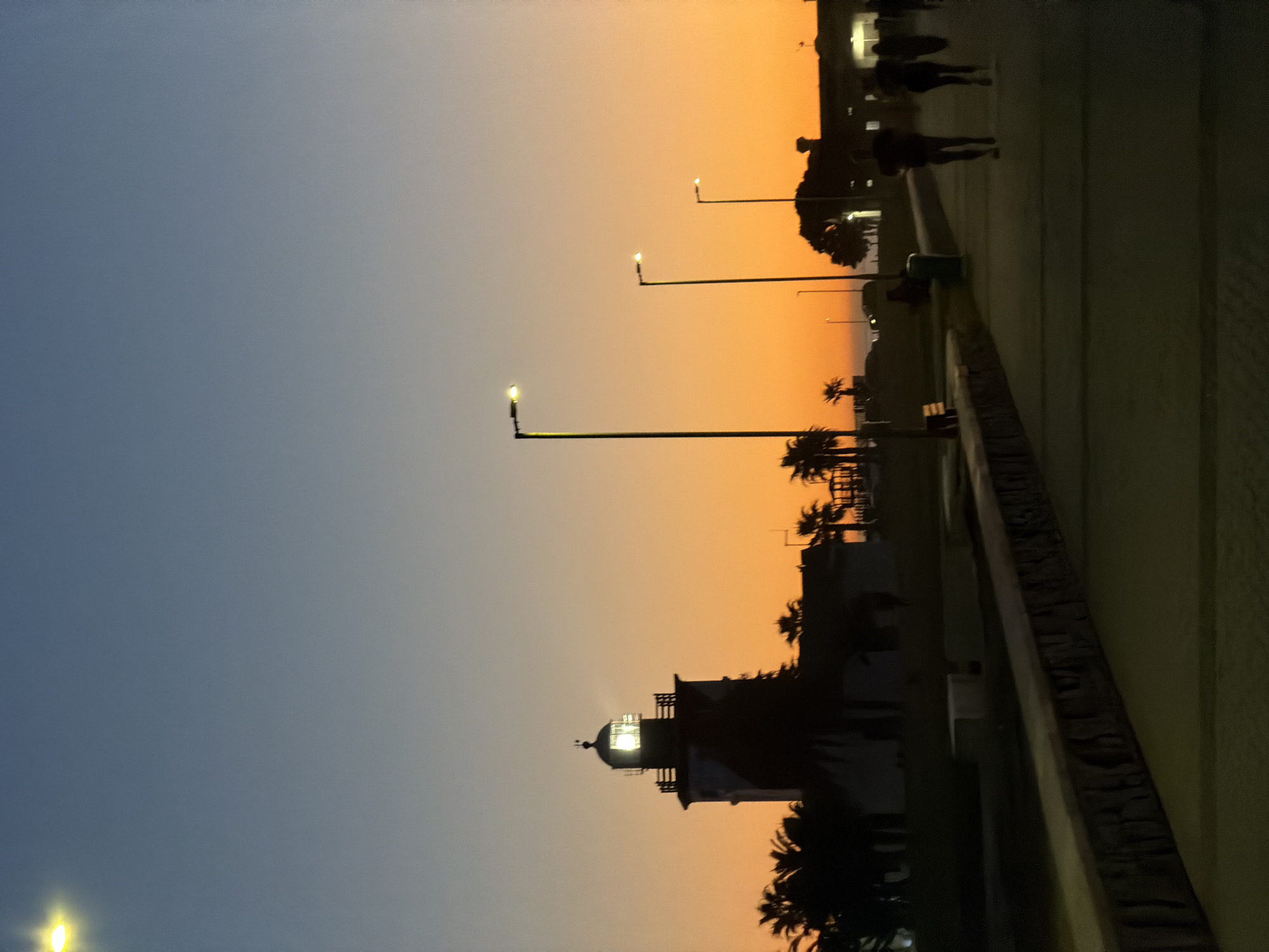 The image shows a street scene at sunset, possibly in Cape Town, South Africa. The sky is a gradient of blue to orange. A lighthouse and palm trees are silhouetted against the sky. Streetlights illuminate the walkway, where people are walking. A low wall runs along the side of the walkway.
