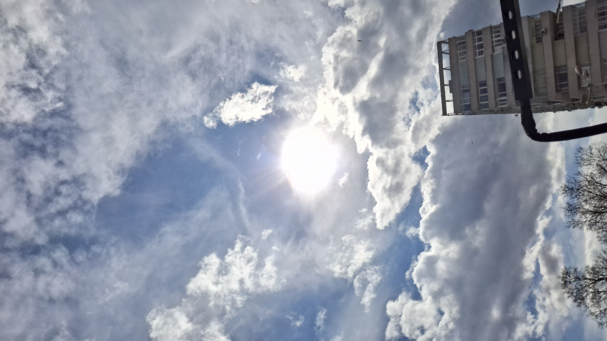 A low-angle shot under bright sunlight, reveals a sky with blue patches interspersed with white clouds. A building is visible on the upper-right, featuring several windows and what appears to be an antenna. Part of a metallic structure juts into the scene from the top, possibly a basketball hoop or similar structure. Bare tree branches are seen on the right. The city location in Varna, Bulgaria, is implied by the architecture and typical weather conditions. No people are visible.