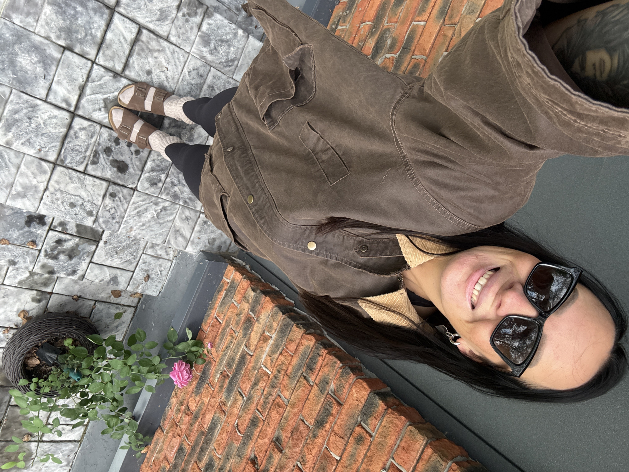 A selfie taken from a high angle shows a woman with long dark hair, wearing black sunglasses, a brown jacket, black leggings, and brown sandals. She is smiling at the camera. The background features a brick wall, a potted rose bush with a single pink bloom, and a tiled outdoor area. The overall impression is that of a casual outdoor setting. The location is not specified as Chaikovsky, Russia, and there is no information in the image to support this claim.