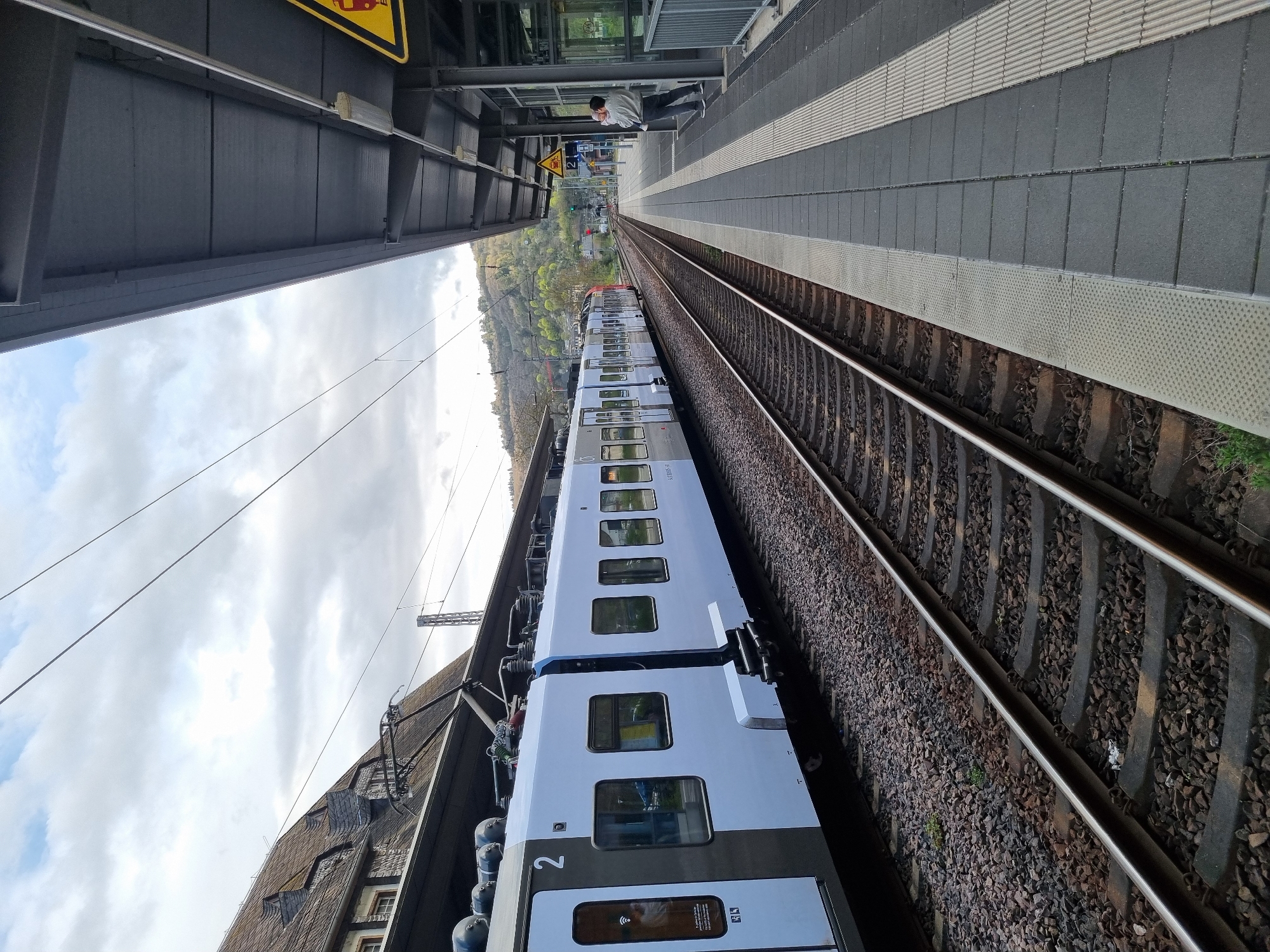 The image depicts a train at the Cochem (Mosel) train station in Cochem, Germany. A white passenger train is stationary on the tracks alongside the platform. A person is standing on the platform. There is vegetation in the background. The sky is overcast.
