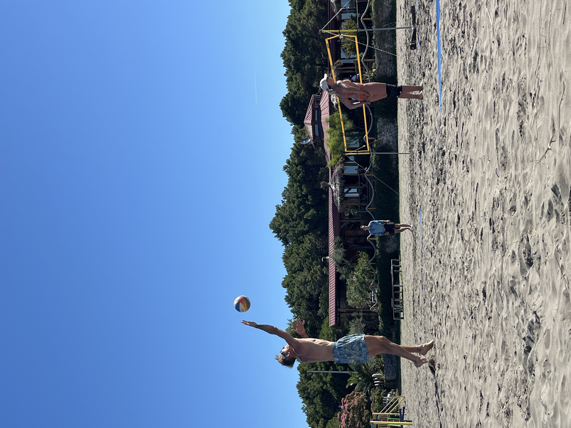 In Pistula, Montenegro, under a clear blue sky, a man in patterned shorts is captured mid-jump on a sandy beach, his arm extended upwards towards a volleyball. A woman in an orange bikini and knee pads stands near a volleyball net, facing away from the camera. In the background, several people are visible near a building with a red-tiled roof, surrounded by lush green trees. The beach is composed of coarse sand with footprints and scattered debris. The scene appears to be a recreational area, possibly a resort, with volleyball courts and seating areas.