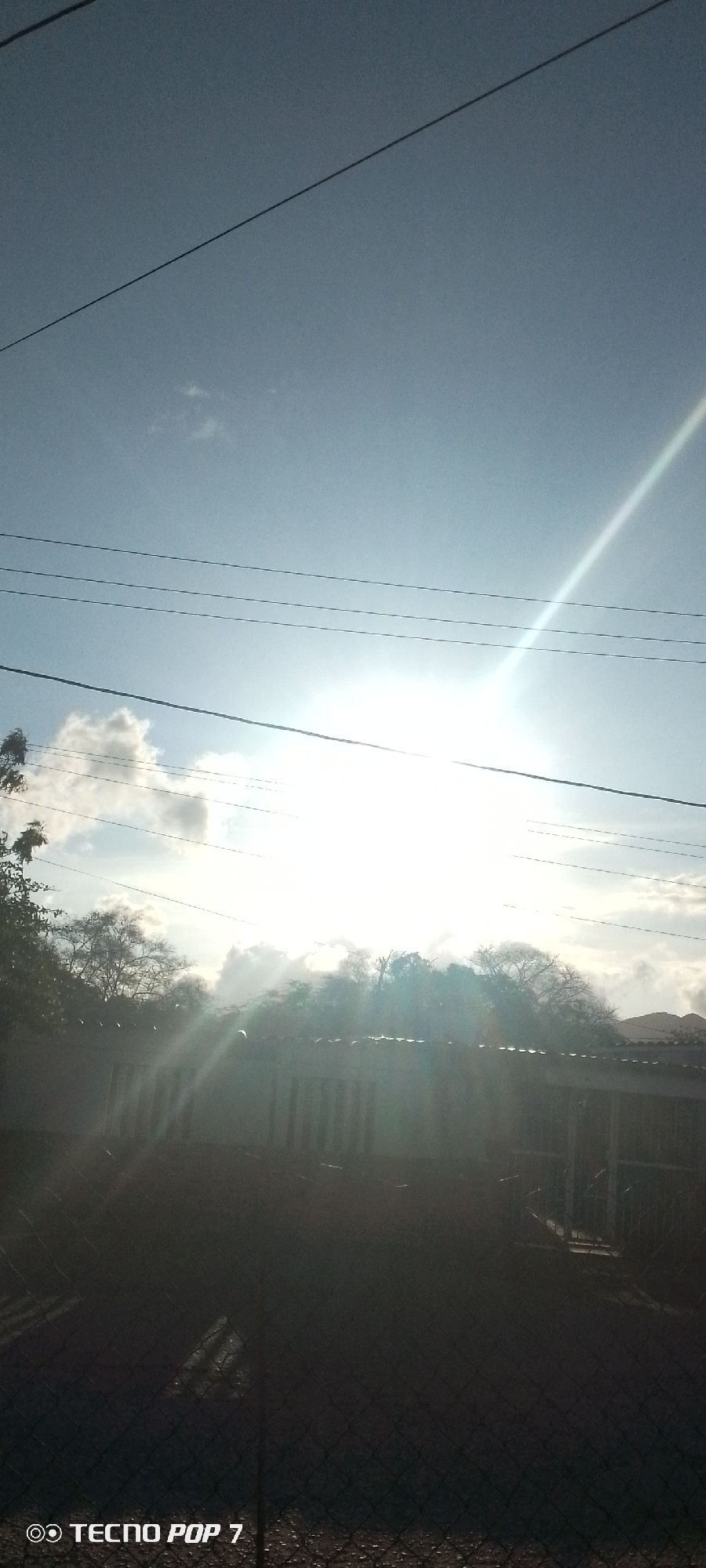 The image shows a bright, sunny day in Altagracia, Venezuela. The sun is high in the sky, partially obscured by thin clouds. There are power lines visible in the upper part of the image. Below, the sun's rays shine through trees and what appears to be a fenced structure or building. The foreground is dark, possibly a shadow cast by the fence. The "TECNO POP 7" watermark is visible in the bottom left corner.