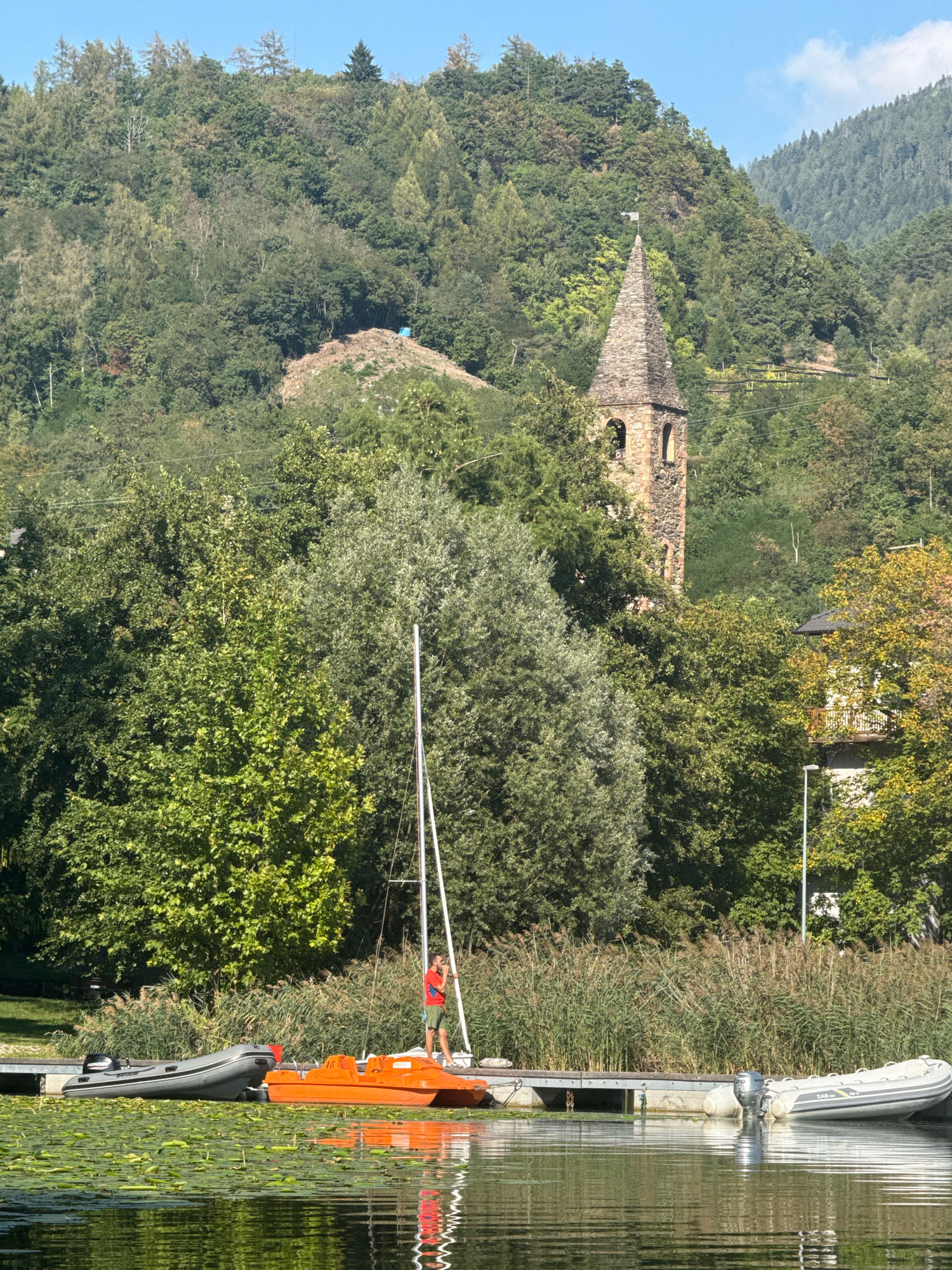 In Pergine Valsugana, Italy, a person in a red shirt and green shorts stands on a wooden dock next to an orange paddle boat and a grey inflatable boat. Behind the dock, tall reeds grow, and further back, a body of water is covered with lily pads. In the background, a stone church tower with a steeple rises above a dense forest that covers a hillside. The sky is clear and blue.