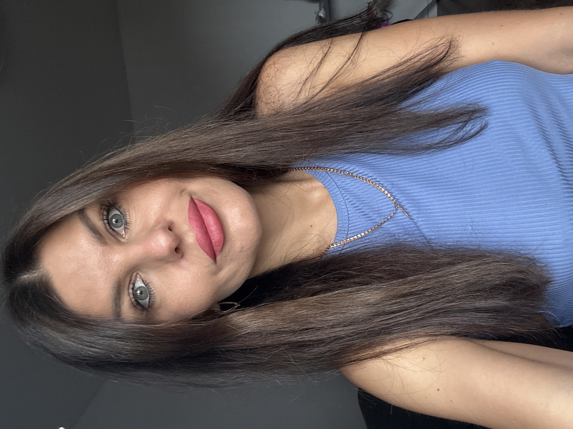 A woman with long, dark brown hair and bright blue eyes is pictured in a close-up, head-and-shoulders shot. She is wearing a light blue, ribbed tank top and a delicate gold necklace. Her lips are painted a vibrant pink. The background is a dark, neutral wall, suggesting an indoor setting. There is no indication of the city of Chaikovsky, Russia, or any specific location.