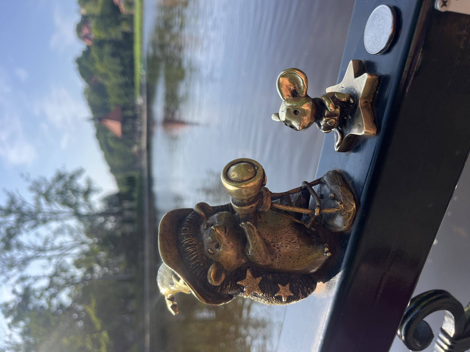 The image shows two bronze figurines on a dark railing next to a body of water. The figurine on the left is a hedgehog wearing a wizard's hat adorned with stars and holding a telescope. The figurine on the right is a mouse holding a camera, standing on a golden star. A silver coin is placed to the right of the mouse. In the background, a calm body of water reflects the sky and trees. On the far bank of the water, there is a gazebo with a red roof and several buildings nestled among green trees under a clear blue sky. This scene is located in Svetlogorsk, Russia, a resort town known for its amber.