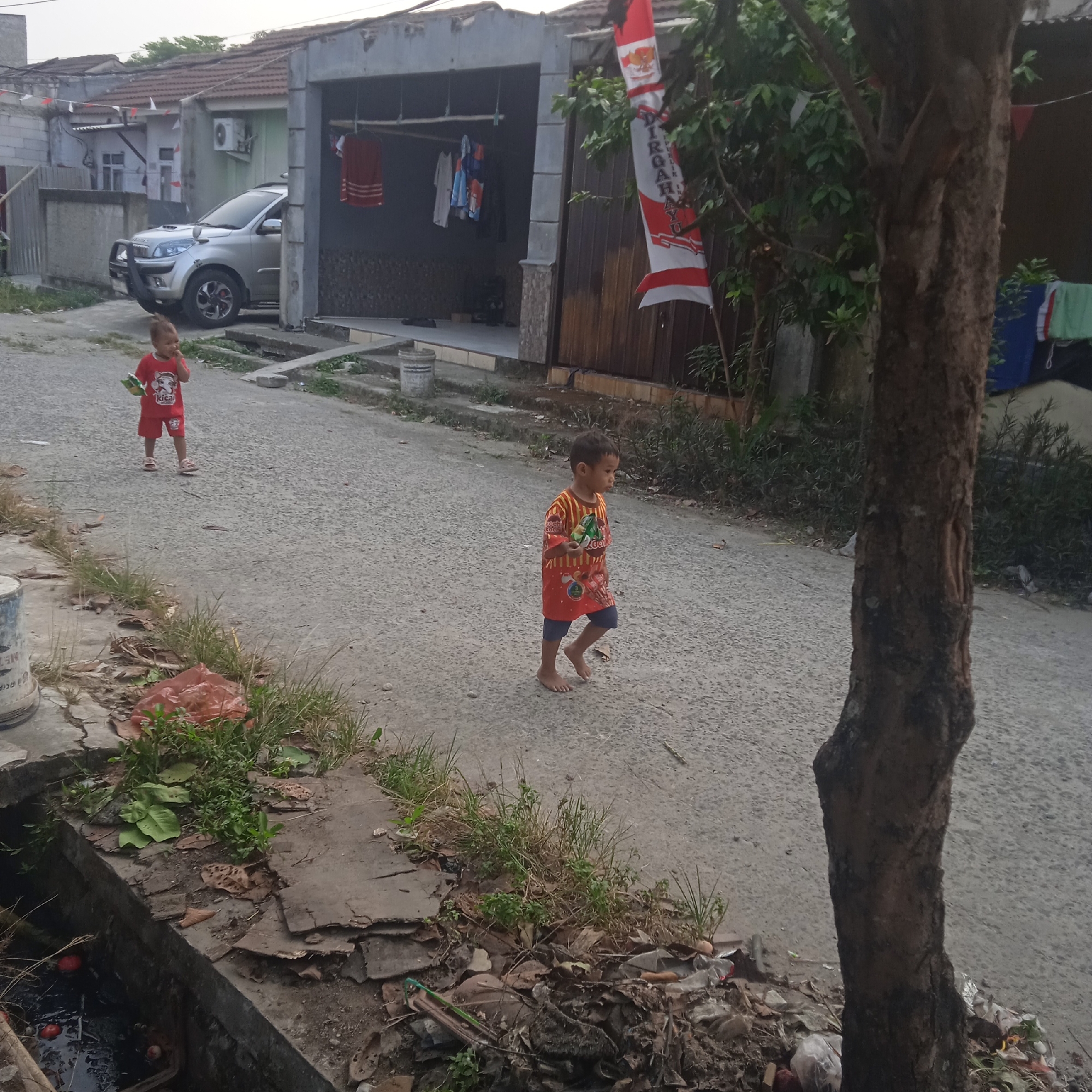 In Situterate, Indonesia, two young children walk along a paved street. The child on the left, wearing a red shirt and red shorts, holds a green object. The child on the right, in a red and orange patterned shirt and blue shorts, also holds a green object. A silver SUV is parked in front of a house. In the foreground, a tree trunk stands to the right, and to the left, a drainage ditch filled with murky water and some red circular objects is visible. The street is lined with houses and vegetation. A national flag, red and white, hangs vertically on the right side of the frame. Clothes are hanging on a line outside one of the houses.