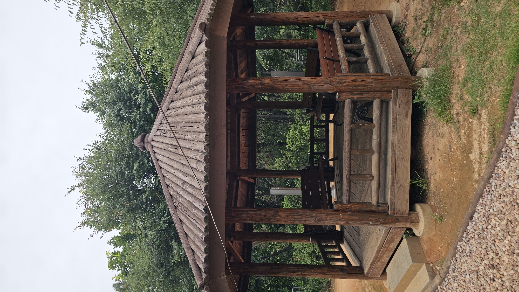 This is a medium shot of an octagonal wooden gazebo located in a park in Asan-si, South Korea. The gazebo has a multi-tiered wooden roof with a rounded finial at the peak. The walls are open, supported by thick wooden posts, and feature wooden benches along the perimeter. The floor of the gazebo is also made of wood. In front of the gazebo, there are stone steps leading up to it, and the ground is a mix of dirt, gravel, and grass. The background is filled with lush green trees. The sky is overcast. There are no people or animals visible in the image.