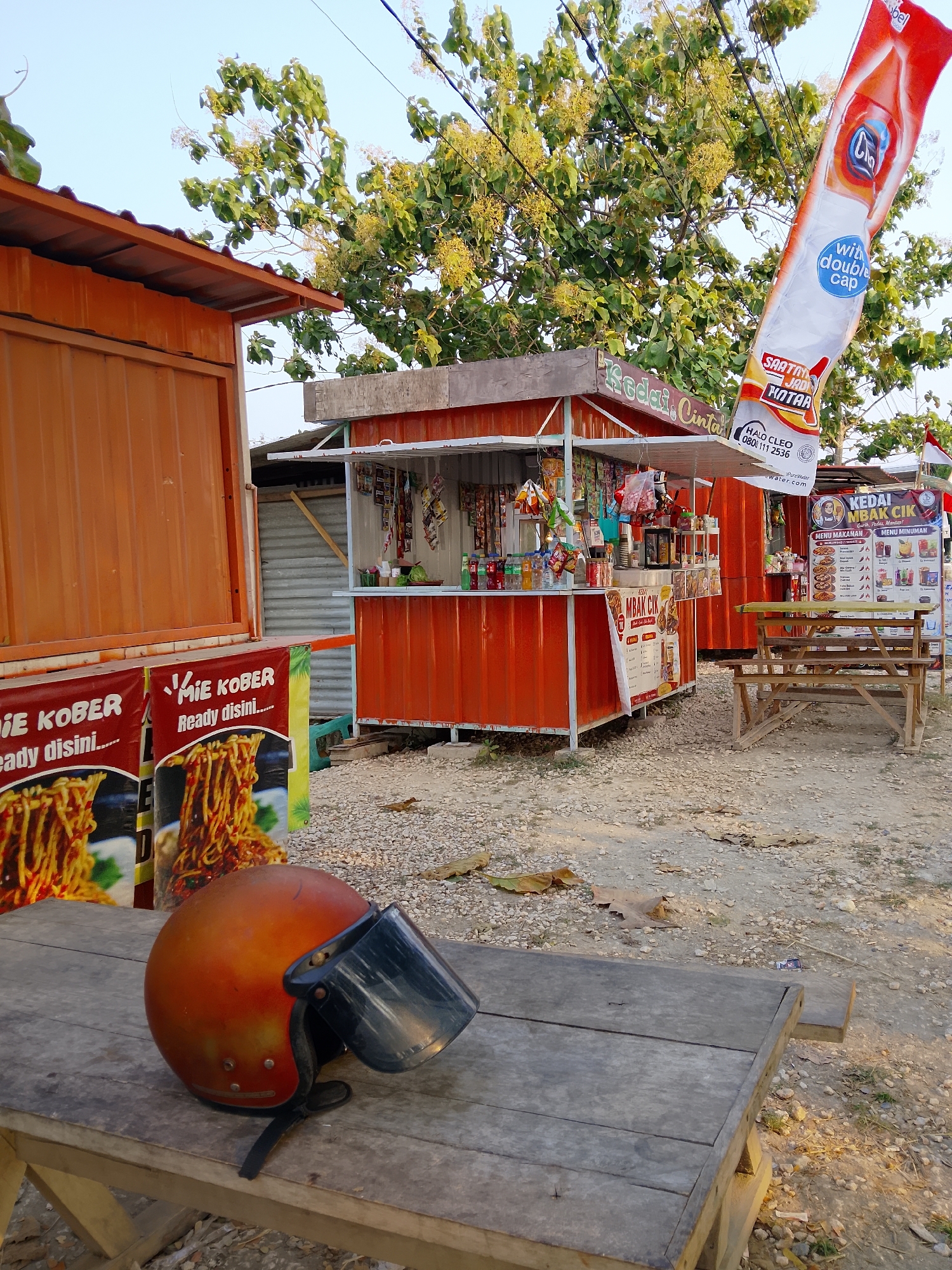 A weathered wooden table in the foreground of the image holds an orange motorcycle helmet with a clear visor. The table is set on a gravelly surface. Behind the table are two orange food stalls, likely shipping containers converted into businesses. The stall on the left displays a banner advertising "Mie Kober," showing an image of noodles. The stall on the right has a menu board displaying food and drink options, and appears to be selling various packaged goods and drinks from an open window. A tall, white and red flag or banner with Indonesian text and an image of a drink bottle hangs from a pole near the right stall. Trees with green leaves and some yellowing blossoms are visible in the background, with power lines crisscrossing the sky. The overall scene suggests a roadside or open-air food court in a rural or semi-urban area, possibly Ngadrin, Indonesia, given the context. The lighting indicates it is daytime, likely late afternoon. There are no people visible in the image.