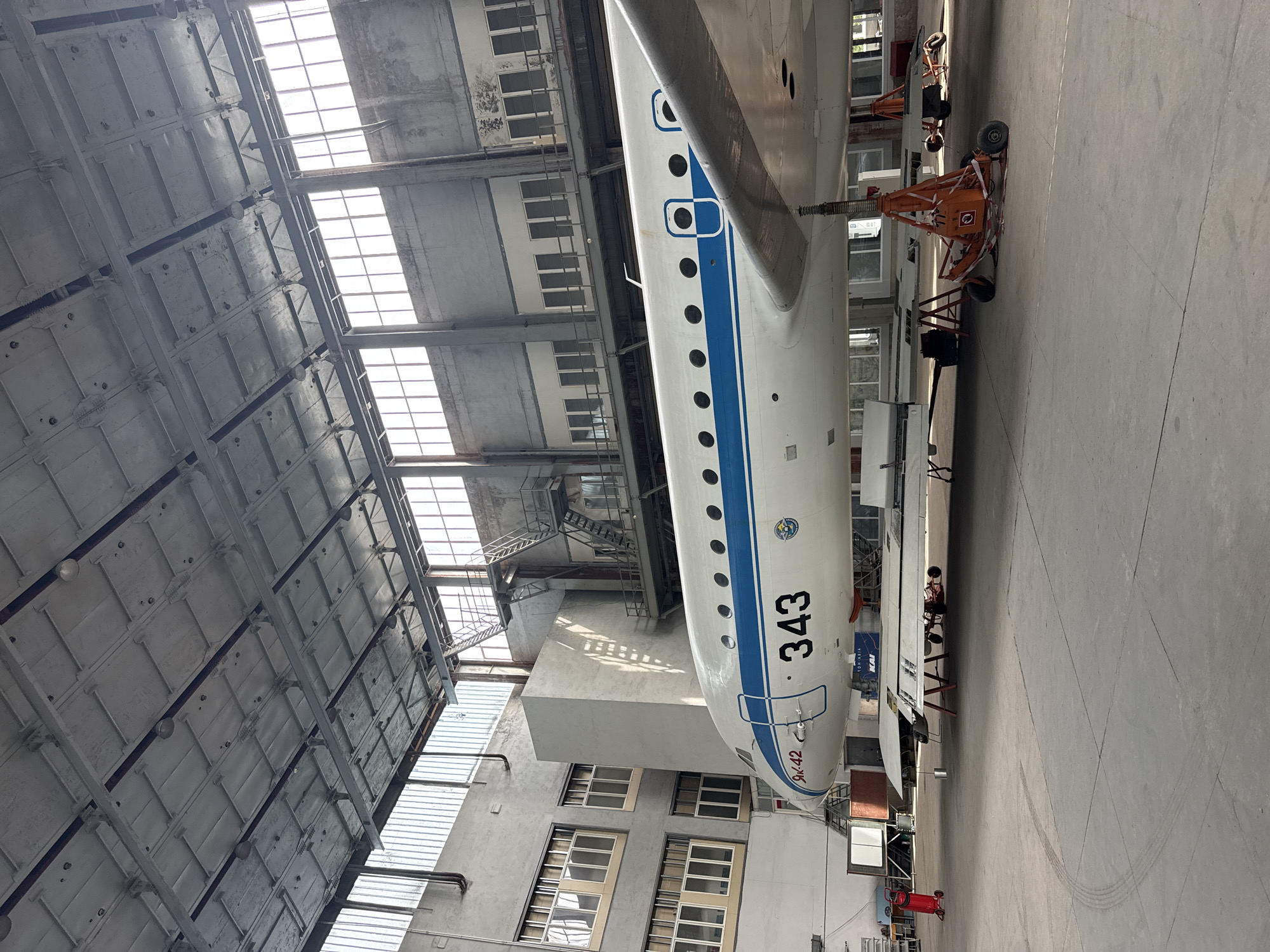 The image depicts the interior of a large aircraft hangar in Kyiv, Ukraine, likely a museum or restoration facility. A white and blue jet aircraft, marked with the number "343" and "ЯК-42", is the central subject, supported by orange jack stands. The aircraft appears to be on display or undergoing maintenance. The hangar itself has a high, industrial ceiling with exposed beams and lighting fixtures. Large windows line the upper walls, allowing natural light to filter in, and concrete walls form the structure. The floor is concrete with tire marks visible in places. To the left of the aircraft, a red fire extinguisher on wheels is parked near some tables and equipment. The overall scene is one of stillness, with no people actively engaged in any interaction within the frame.
