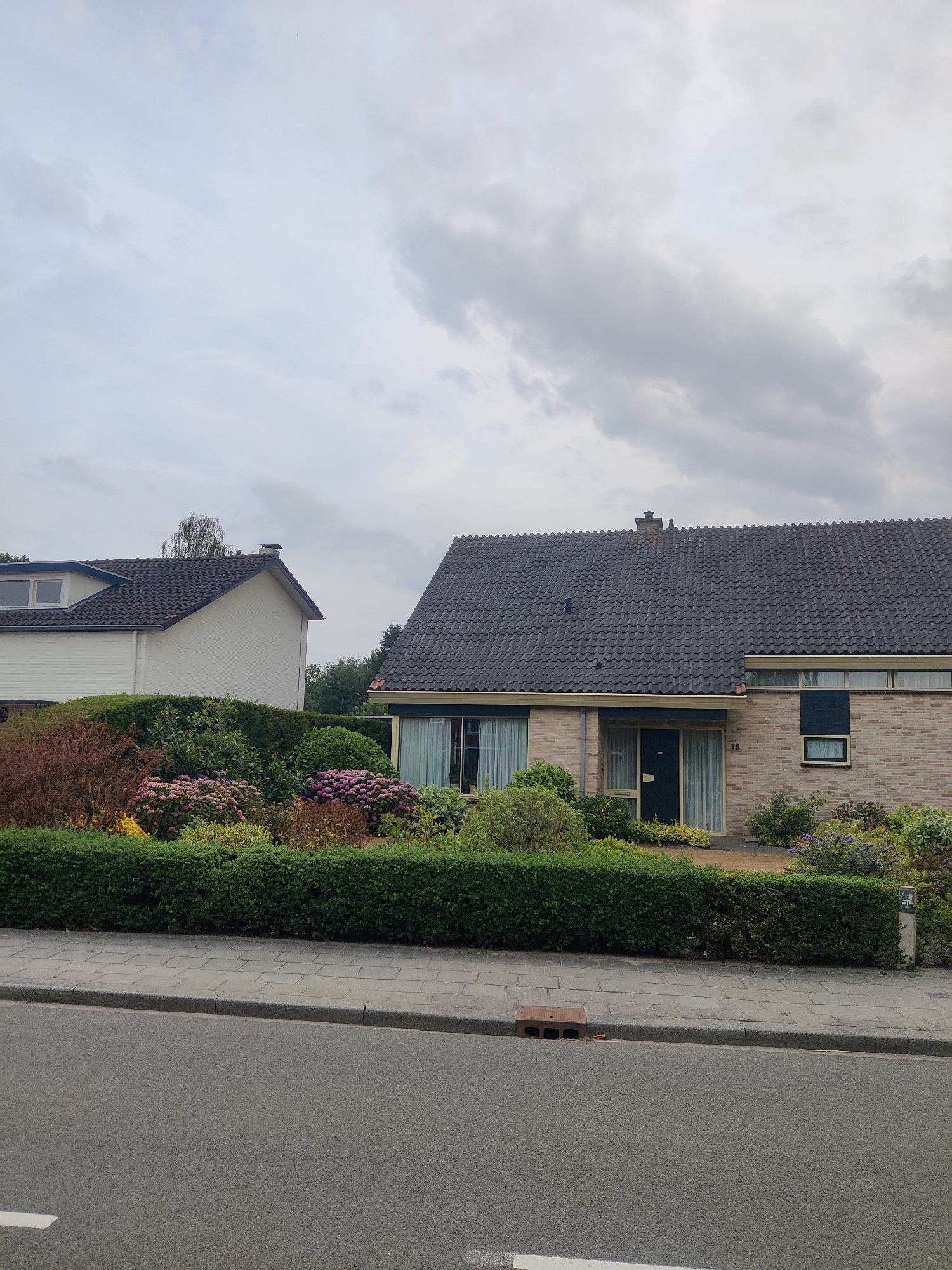 The image depicts a residential street scene in Winterswijk, Netherlands. The foreground shows an asphalt road with a gray tiled sidewalk and a low green hedge separating it from a well-maintained garden. Behind the hedge, two houses are visible. The house on the right is a single-story brick building with a dark tiled roof, featuring a dark blue door, several windows, and a small numerical sign "76". The house on the left is a larger, white two-story structure with a dark tiled roof and dormer windows. The garden area is lush with various bushes and flowering plants in shades of pink, red, and yellow. The sky is overcast with gray clouds. The overall impression is one of a quiet, suburban environment.