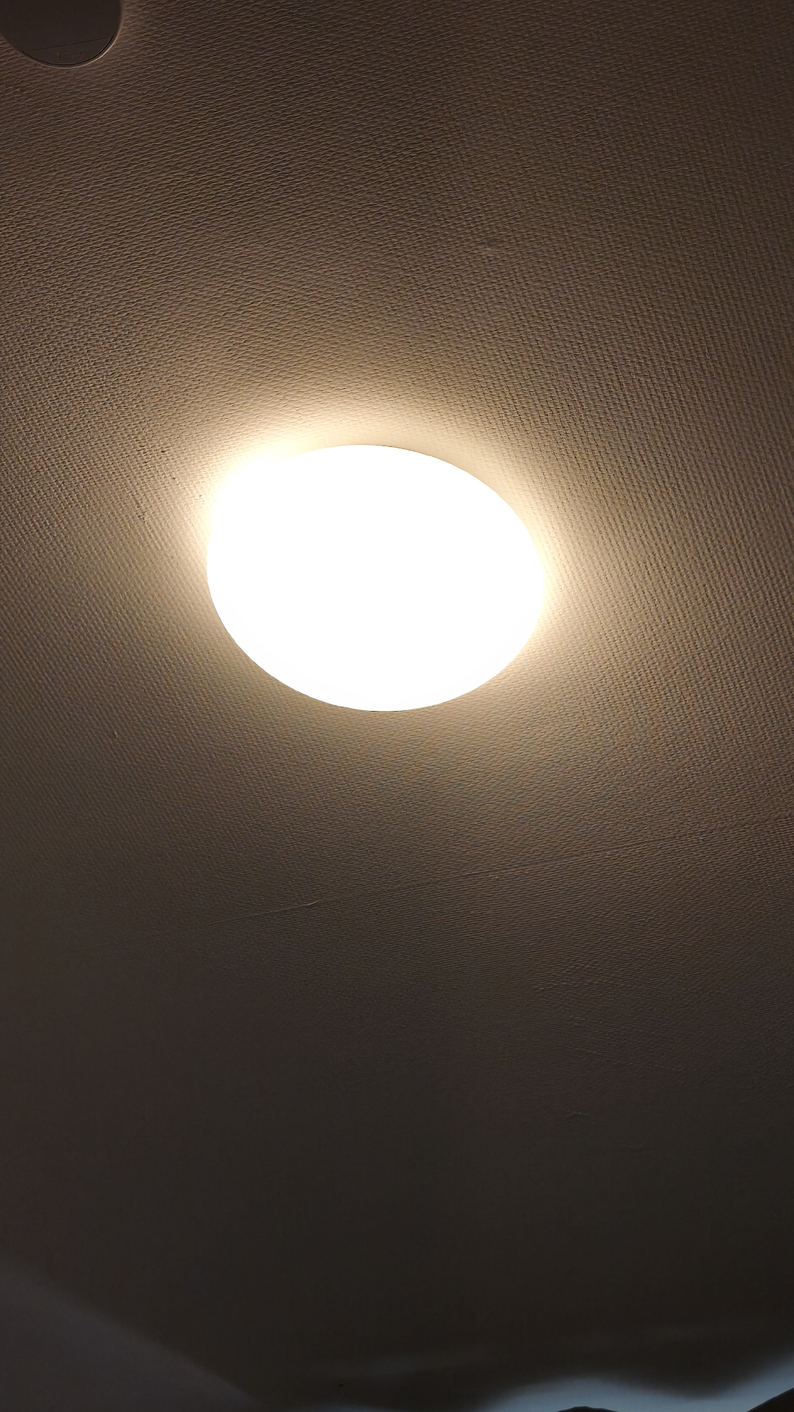 The image shows a ceiling-mounted, round light fixture illuminating a textured ceiling. The light is centrally located in the frame and is the brightest element. The ceiling appears to be a light neutral color. The location is unknown.