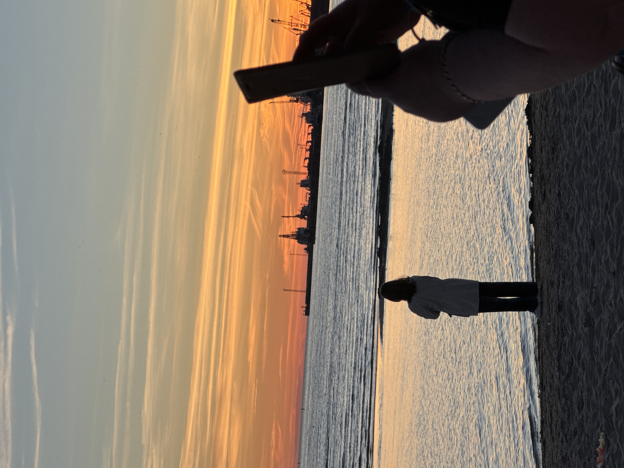 The image depicts a sunset scene at a beach in Tallinn, Estonia. In the foreground, a person is taking a photo with a smartphone. In the midground, a solitary figure stands by the water's edge, facing the sea. The background shows a harbor with cranes and ships silhouetted against the colorful sunset sky. The water is calm with gentle waves lapping at the shore. The sand in the foreground is textured with footprints and some debris.