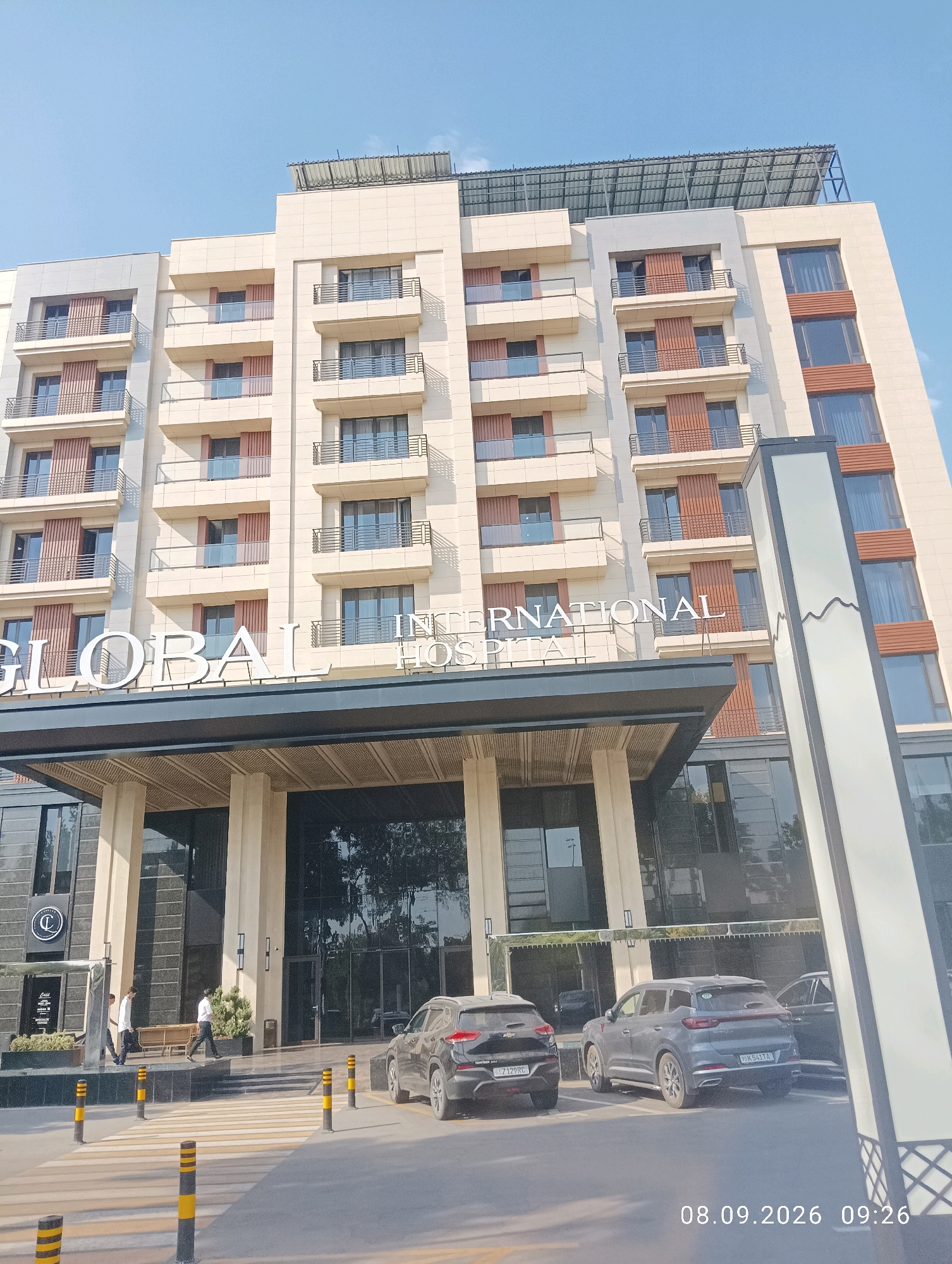 The image shows the exterior of the Global International Hospital in Tashkent, Uzbekistan. The modern building features multiple stories with balconies and large windows. In the foreground, a crosswalk is visible with yellow and black bollards. Two cars are parked in front of the hospital entrance. Several individuals are walking towards the entrance, dressed in casual and business attire. The sky is clear and blue, suggesting a daytime scene. The timestamp "08.09.2026 09:26" is overlaid on the bottom right of the image.