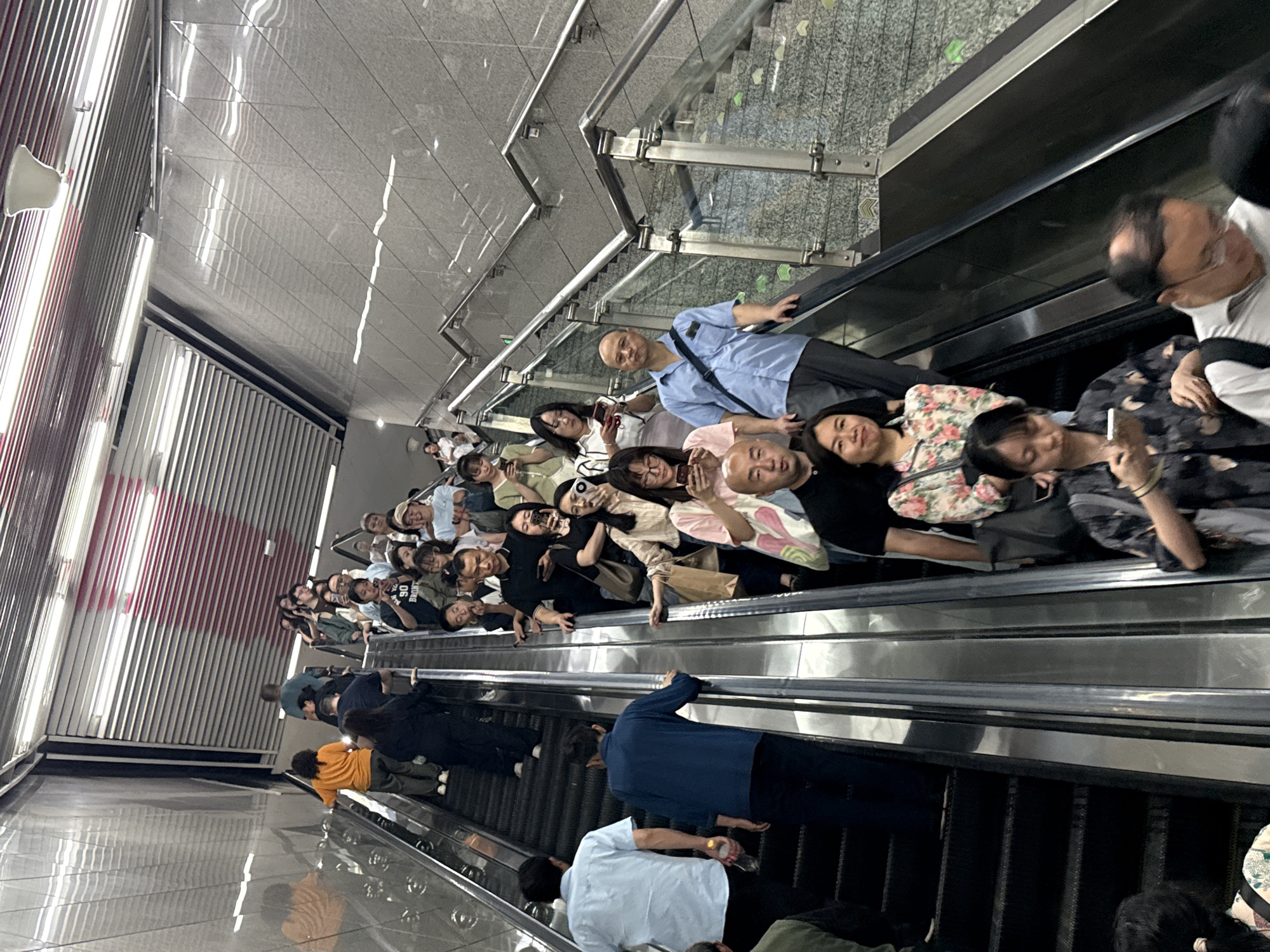 The image shows a large group of people riding an escalator in the Yuzhong District, China. The escalator is going upwards. Most of the people are holding phones. The background has tiled walls and metal railings.