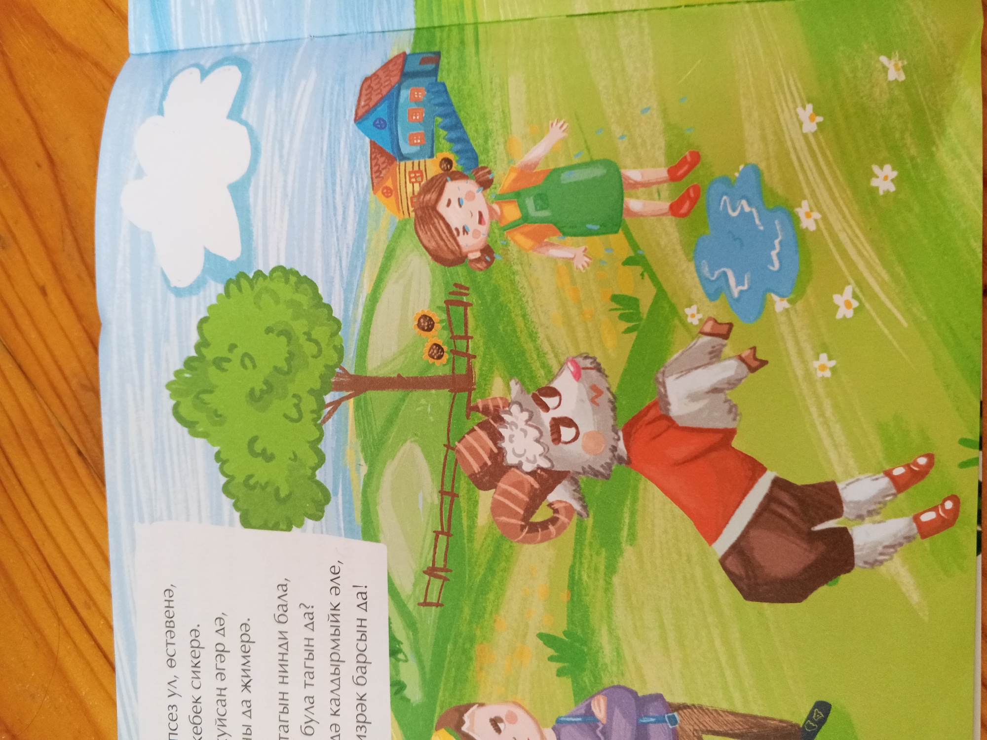 The image depicts a colorful illustration from a children's book, featuring cartoonish characters in an outdoor, rural setting. In the foreground, a young girl with brown pigtails and a green dress is crying, with visible tears. Behind her, a stylized blue puddle suggests recent rain or water. Further back, a goat-like creature with horns and a fluffy face wears a red shirt and brown pants, appearing to be in motion. A small, multi-colored house with a red roof and yellow and blue walls is visible in the upper left background. The environment is a bright green, grassy landscape dotted with small white flowers. The bottom portion of the image contains text in an unknown language, likely a narrative accompanying the illustration. The image itself is part of a larger book, with a wooden surface visible to the left, suggesting it is open on a table. There is no information within the image to indicate its location in Chistopol, Russia, or any other specific geographical place.