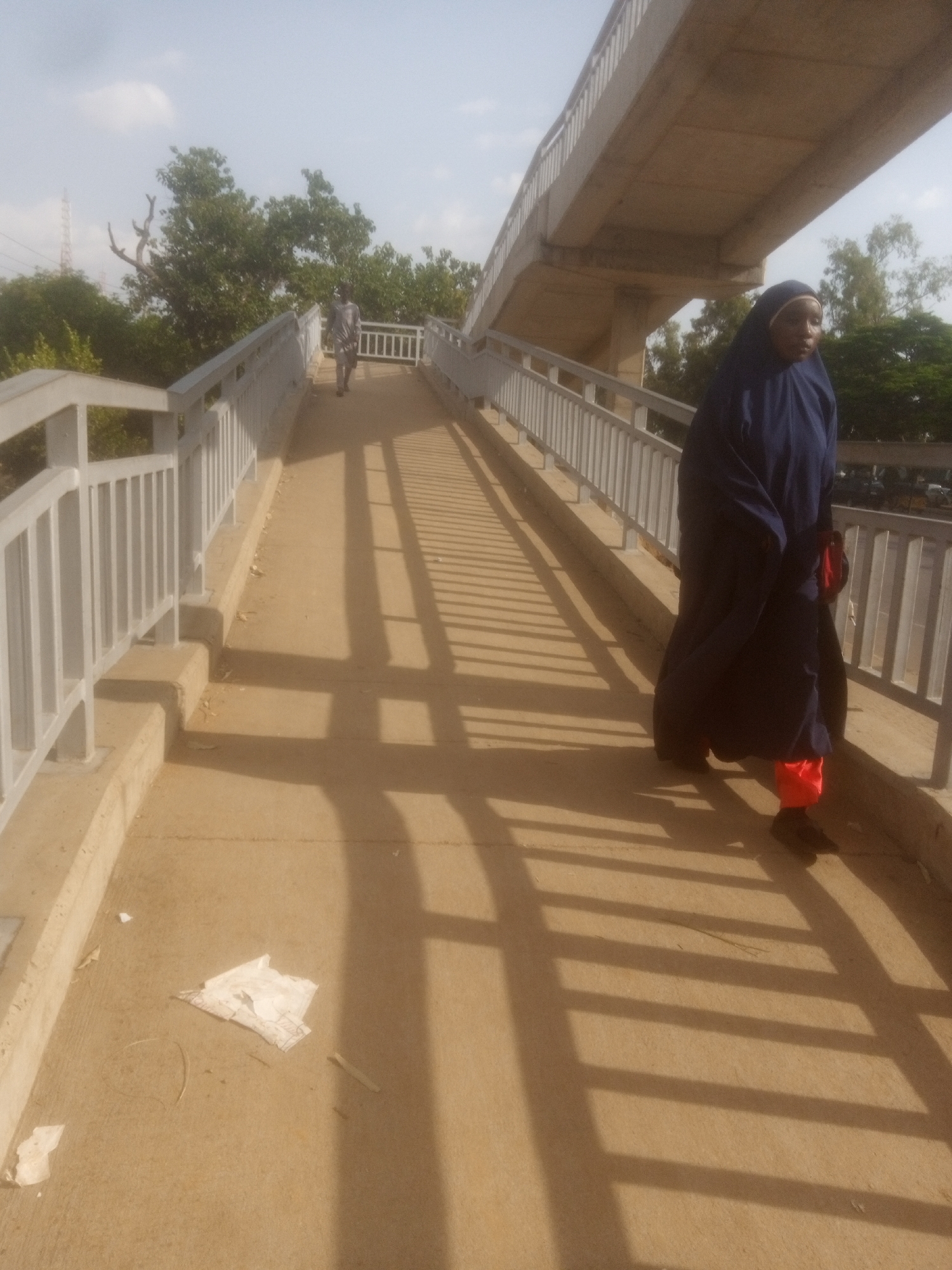 The image depicts a pedestrian overpass in Unguwan Dosa, Nigeria. A woman wearing a dark blue hijab and orange pants is walking towards the viewer on the overpass. Her body is partially obscured by her clothing. In the background, a second individual, dressed in a light-colored garment, is walking away from the viewer further down the overpass. The overpass itself is constructed of concrete with metal railings on either side. The ground surface of the overpass is a light brown, sandy color. Sunlight casts long, distinct shadows of the railings across the walkway. There is some debris, including a crumpled piece of paper, on the overpass surface. Trees and foliage are visible on either side of the overpass, and a power line tower can be seen in the distance. The sky is overcast with some bright clouds.