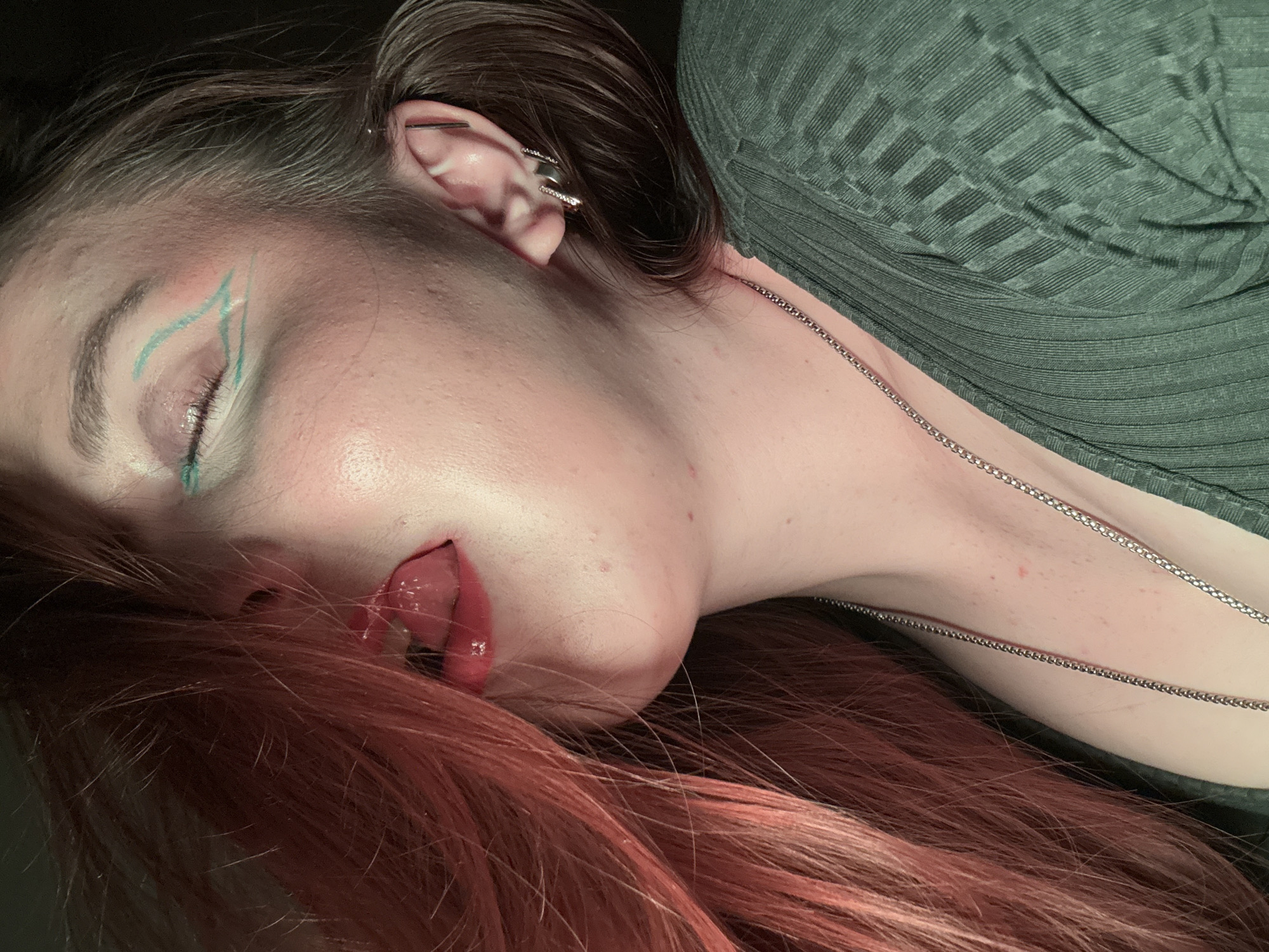A person lies on their side with their eyes closed and lips slightly parted. The person has red-brown hair covering part of their face and silver jewelry in their ear. They wear green eyeliner and a green top with a textured pattern. A silver necklace chain rests on their neck and chest. The setting appears to be indoors. There is no evidence to suggest this image was taken in Izhevsk, Russia.